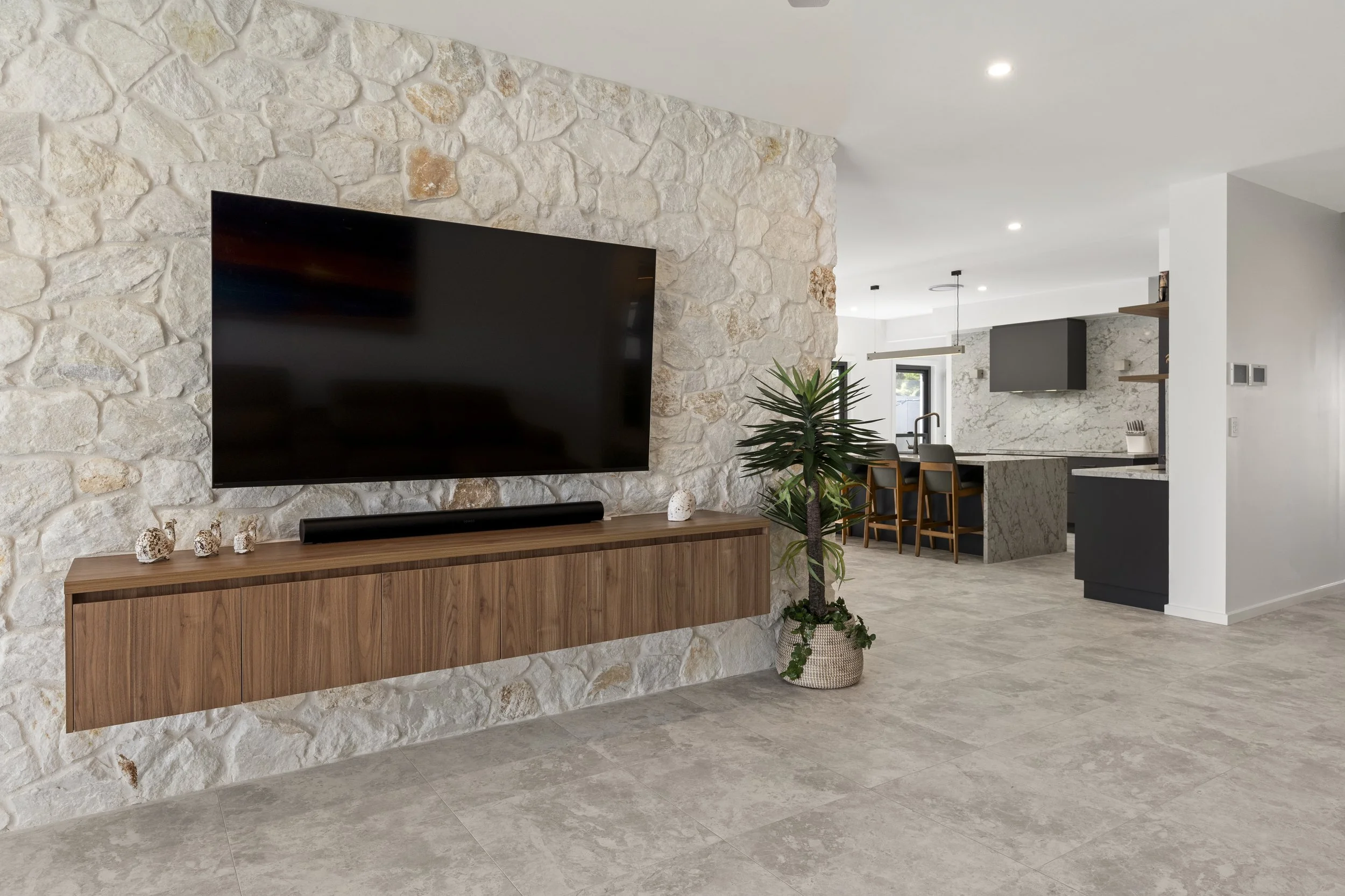 Modern living room with stone accent wall, flat-screen TV, wooden console, painting of seashells, potted plant, and kitchen with bar stools in background.