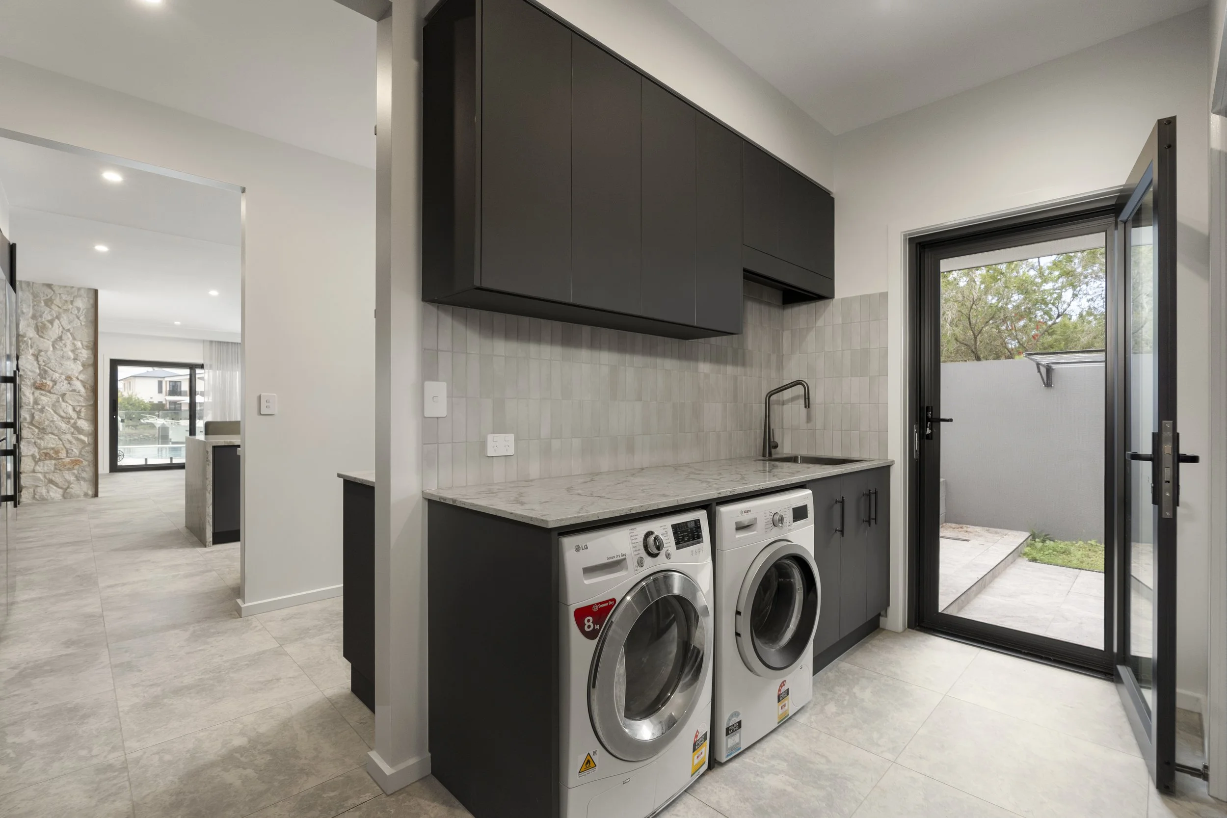 A laundry room with black cabinetry, a stone accent wall, and a view of a patio outside. There are two front-loading washing machines below the counter, and a sink with a faucet. A door opens to an outdoor area with greenery and trees.  Action Kitche