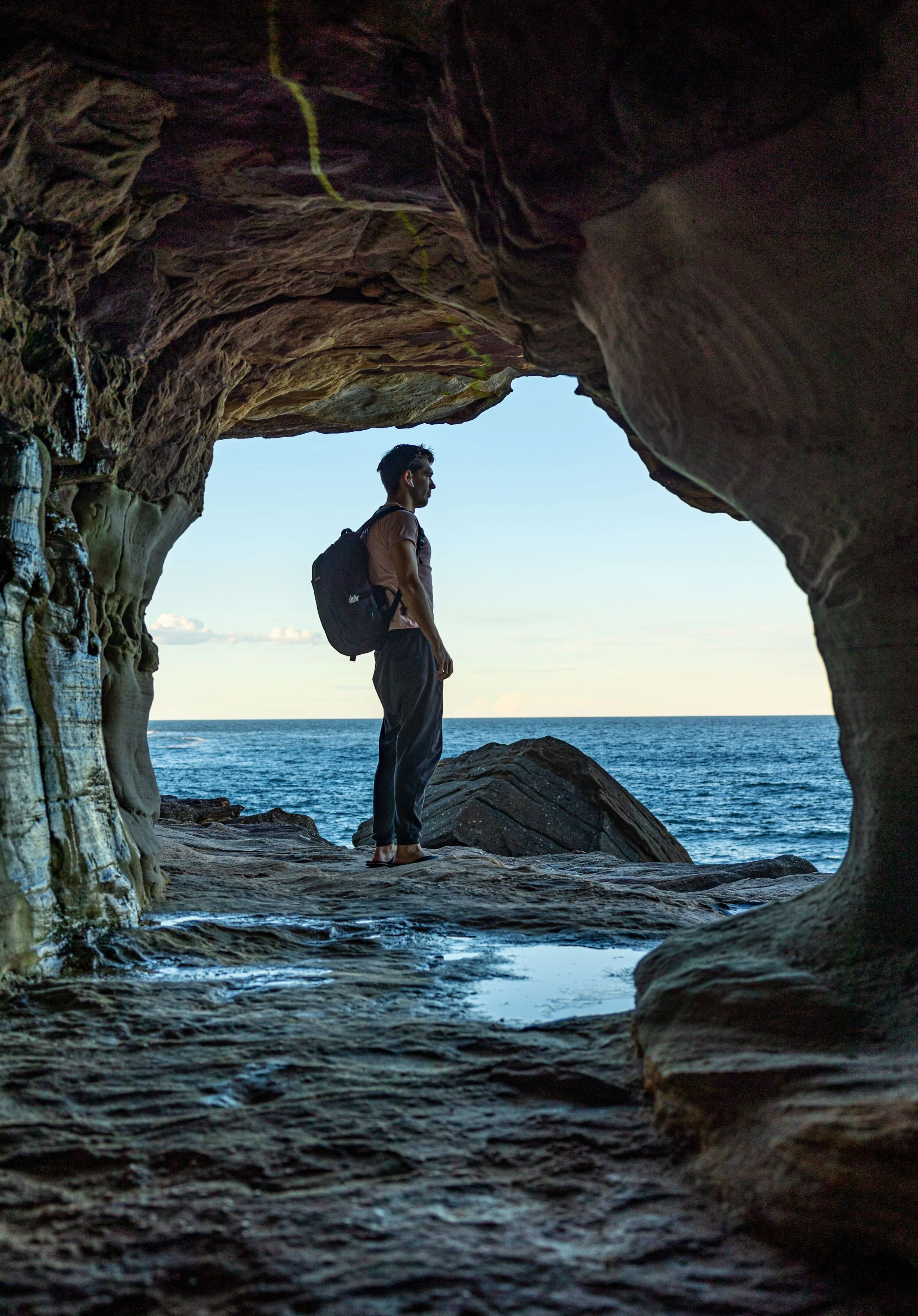 A person with a backpack stands on rocky shore inside a sea cave, looking out at the ocean with clear sky above.