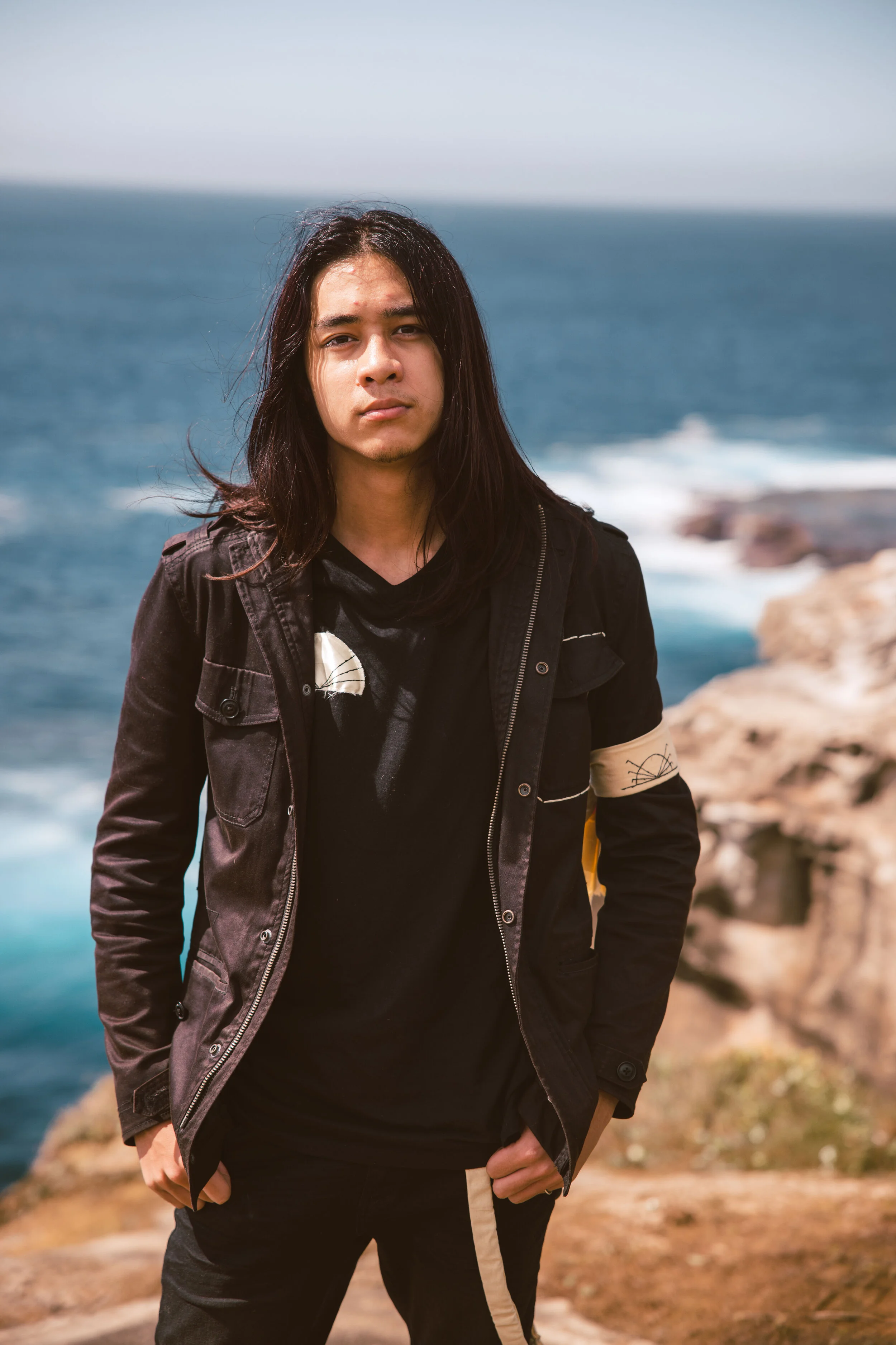 Young man with long dark hair standing outdoors near rocky coastline and ocean in the background.