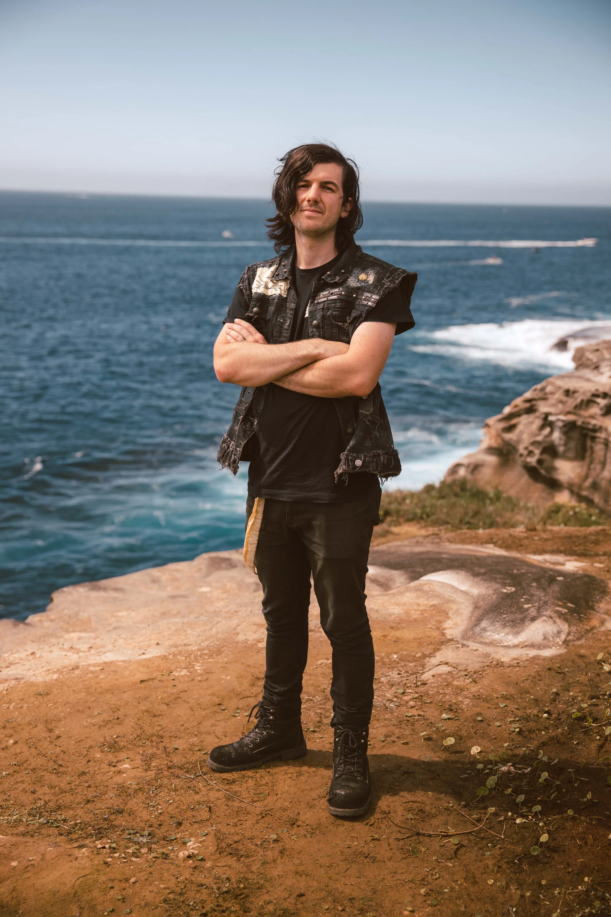 A young man with long dark hair, crossed arms, wearing a sleeveless vest and black boots, stands on a rocky cliff overlooking the ocean under a clear sky.