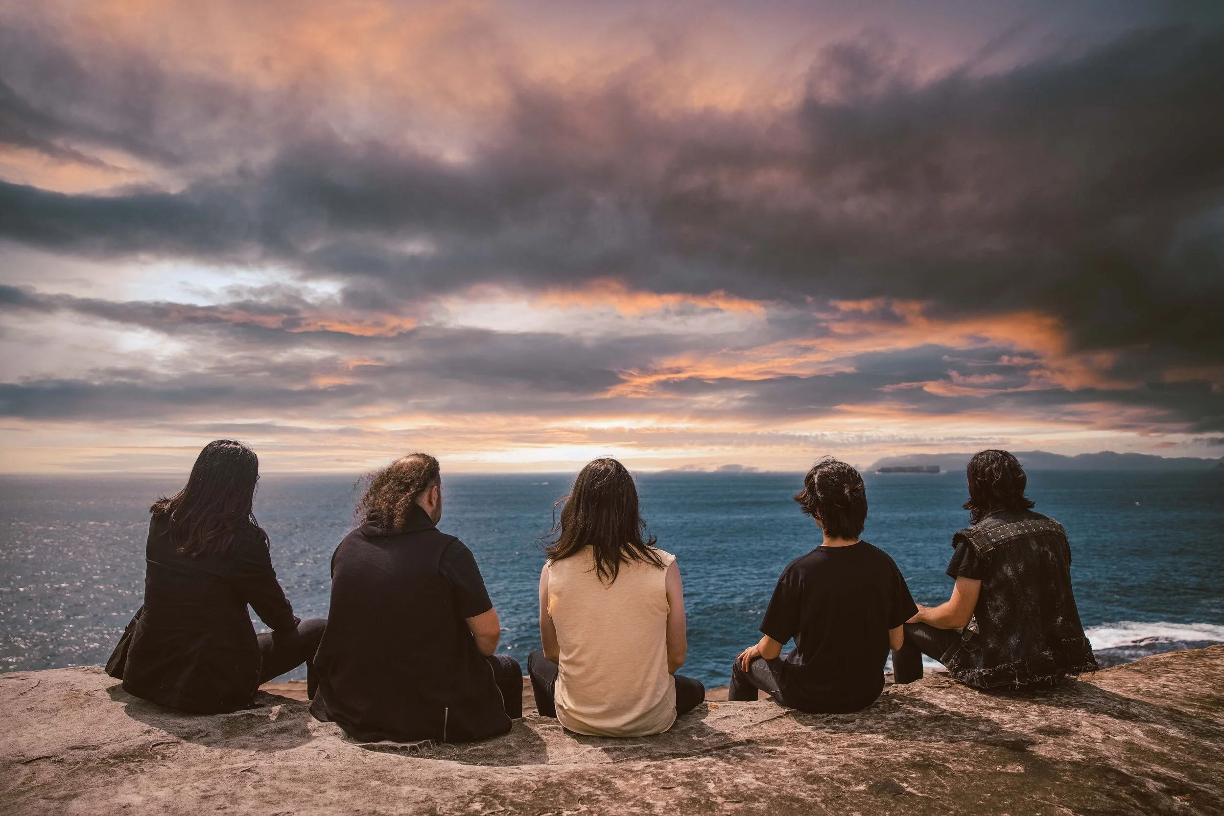 Five people sitting on a rocky ledge watching a sunset over the ocean with clouds in the sky.