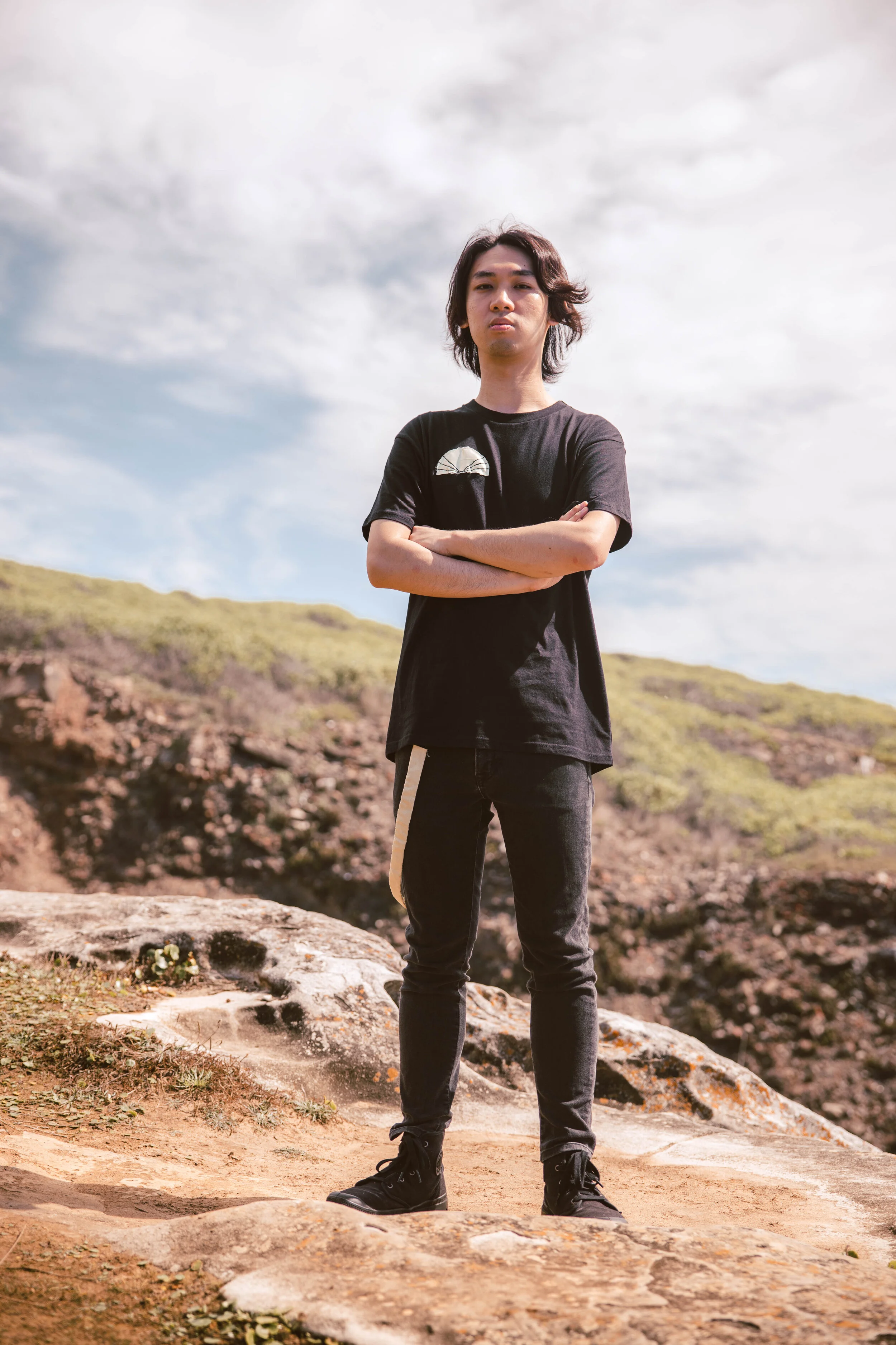 A young man with medium-length dark hair standing outdoors with arms crossed, wearing a black t-shirt and black jeans, against a cloudy sky and a rocky hill in the background.