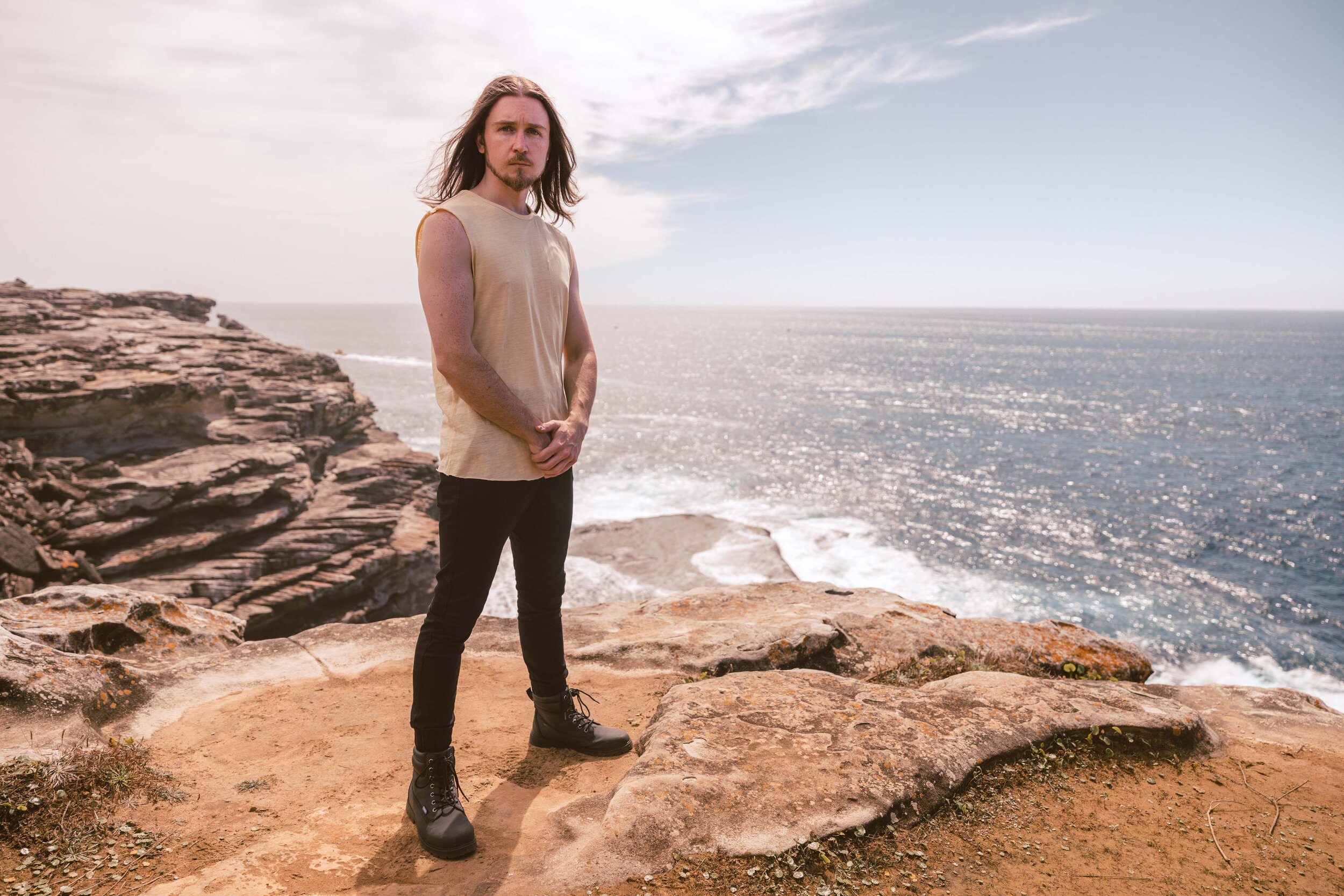 A man with long hair and a beard standing on rocky cliffs overlooking the ocean, wearing a sleeveless beige top, black pants, and hiking boots.