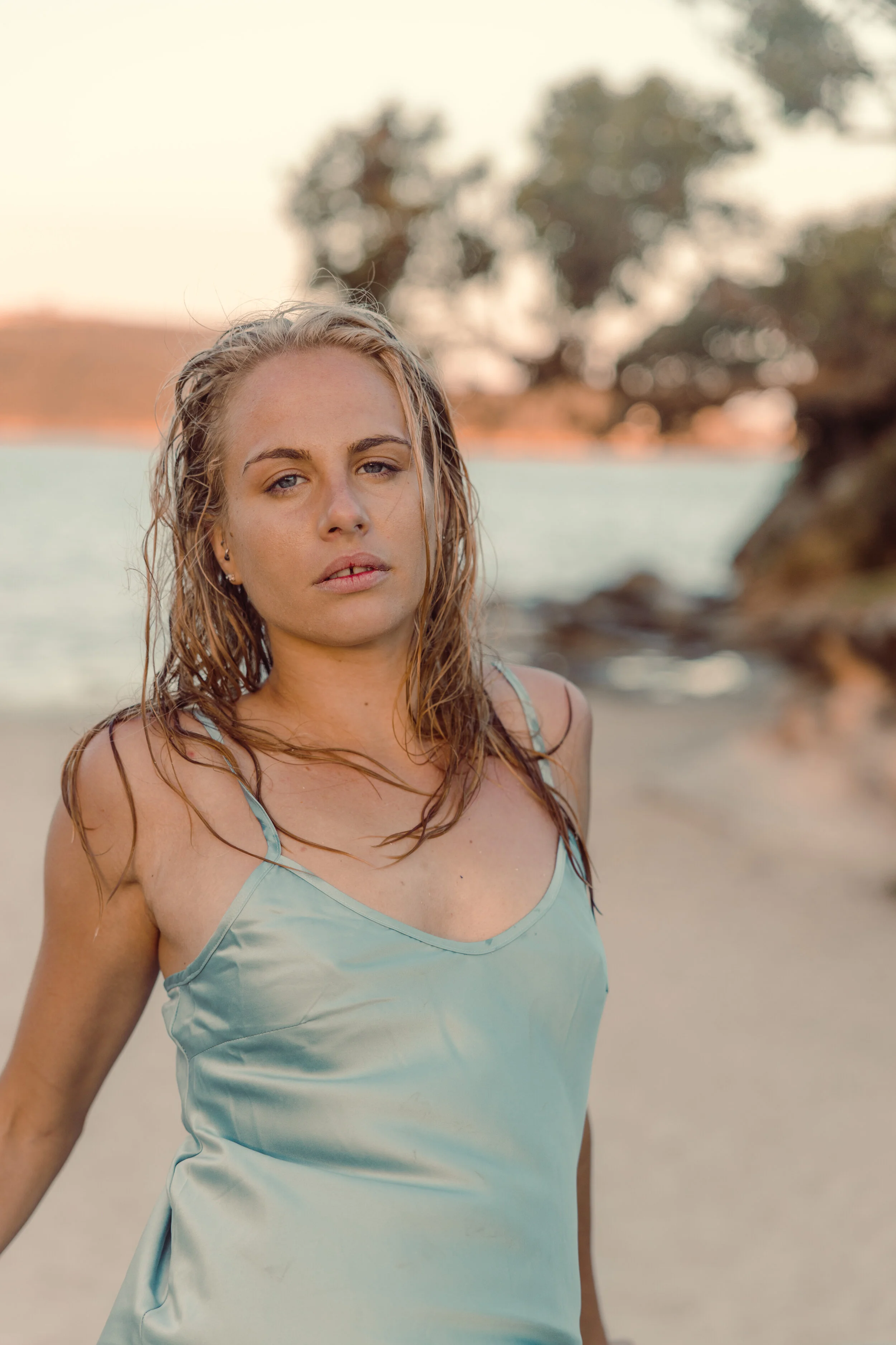 A woman with wet hair wearing a light blue sleeveless top standing on a beach with water, trees, and rocks in the background during sunset.