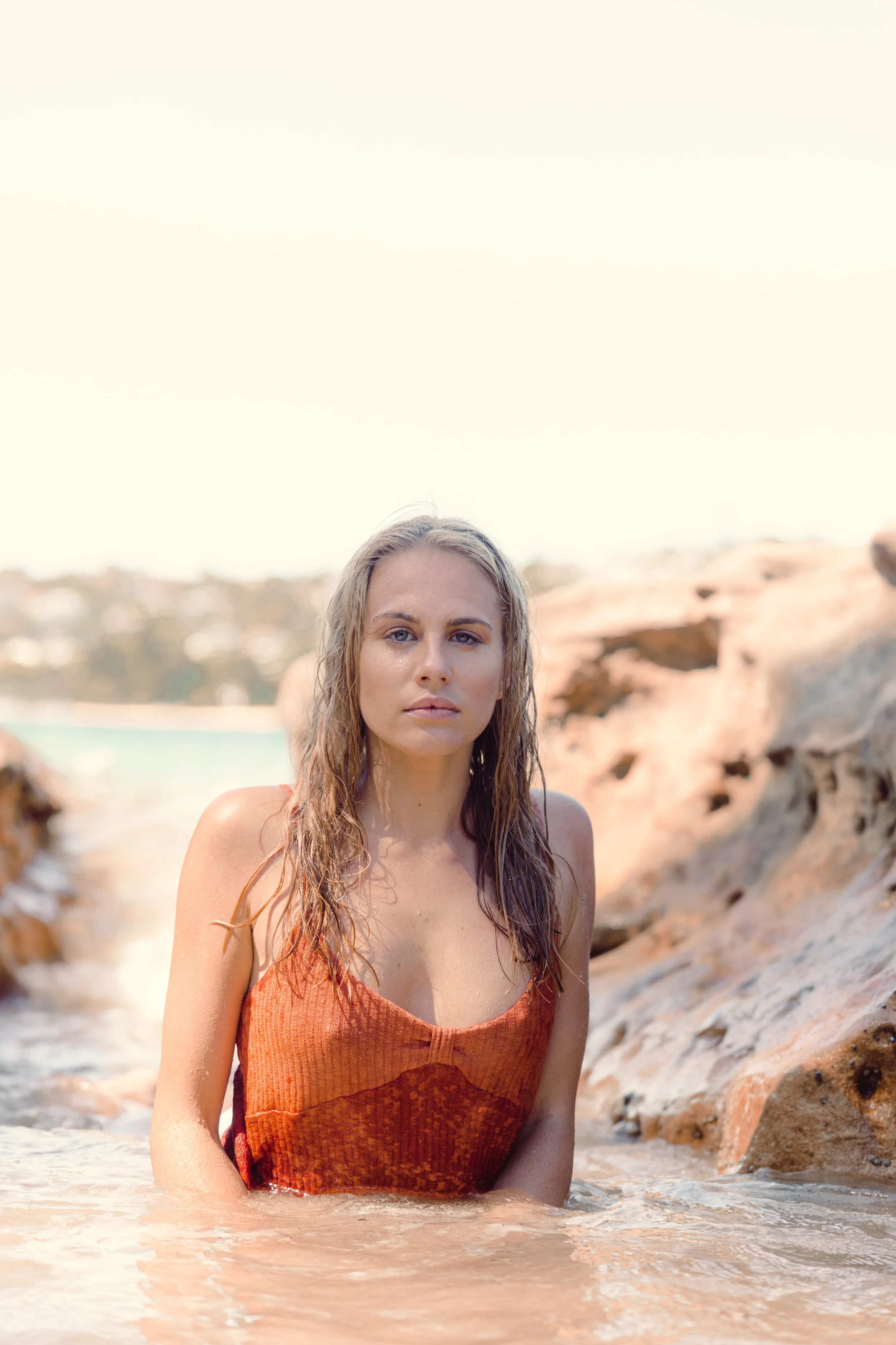 Woman in an orange swimsuit standing in shallow water near rocks at the beach, with a distant coastline in the background.