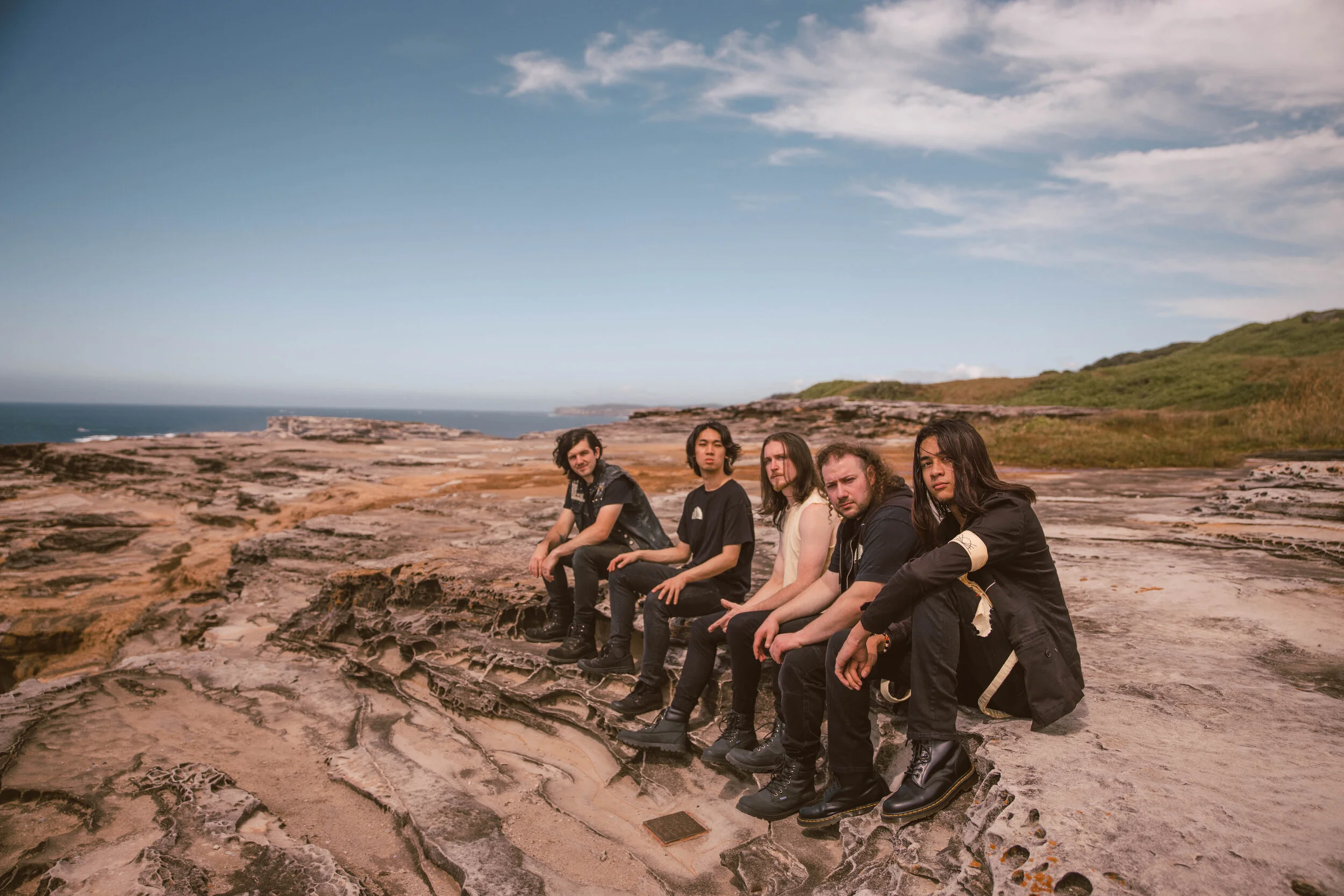 Five young adults sitting on a rocky landscape near the coast, with a blue sky and a few clouds overhead, and grassy hills in the background.