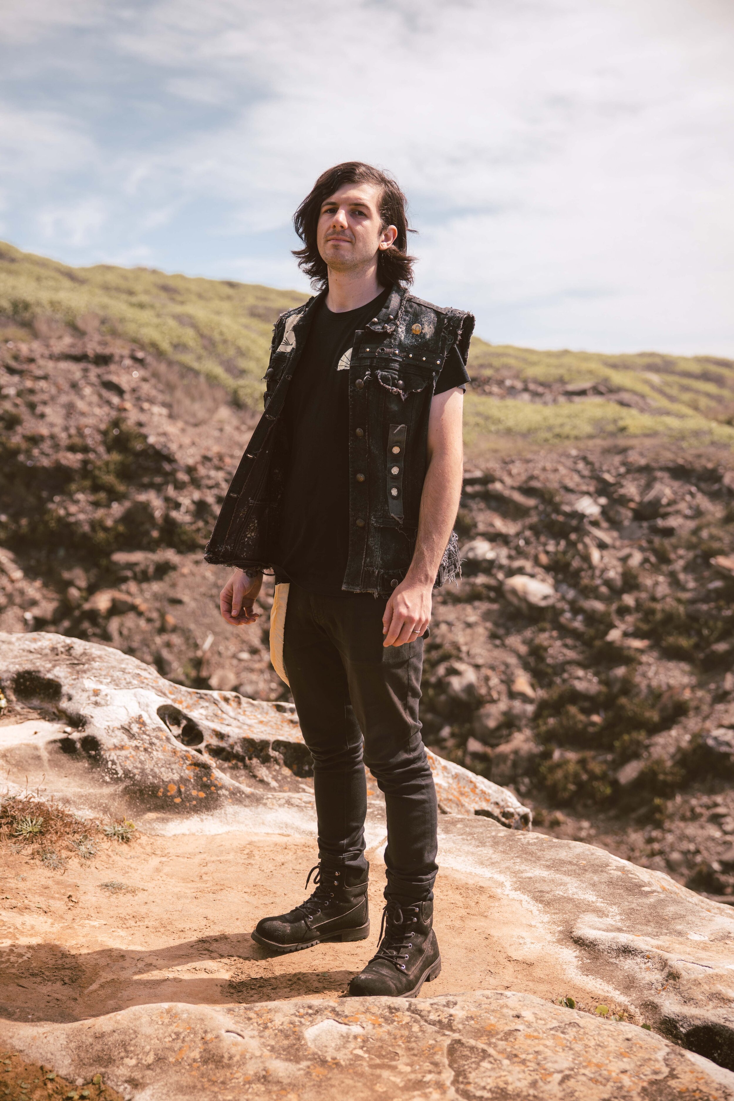 A young man with long, dark hair stands on a rocky terrain outdoors under a partly cloudy sky, wearing a black sleeveless denim jacket, black pants, and black combat boots.