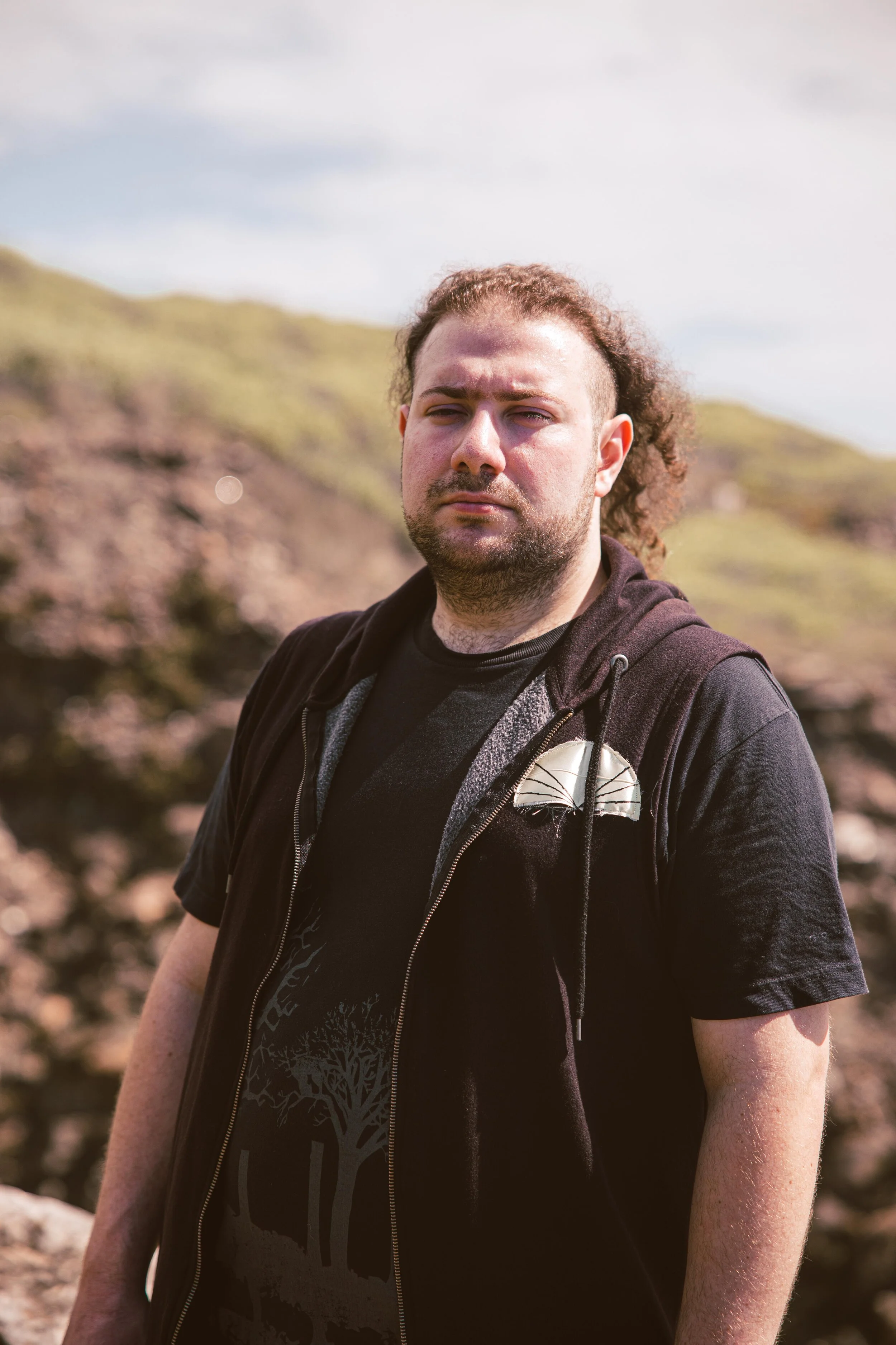 A man standing outdoors in a natural landscape with hills and rocks in the background, wearing a black hoodie and a T-shirt with a tree graphic, looking at the camera.