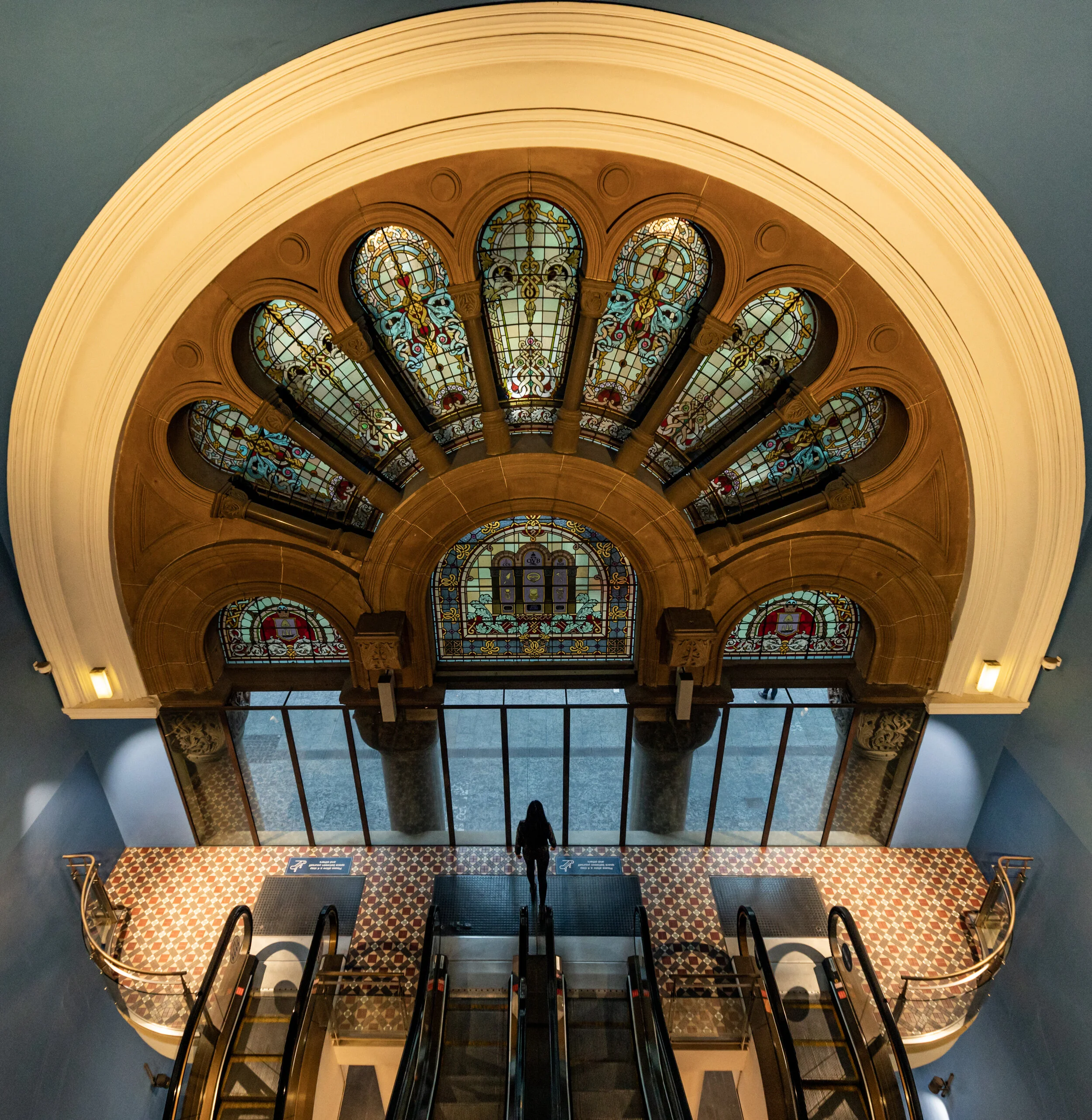Interior view of a building with stained glass windows, an arched ceiling, and a person standing on an escalator.