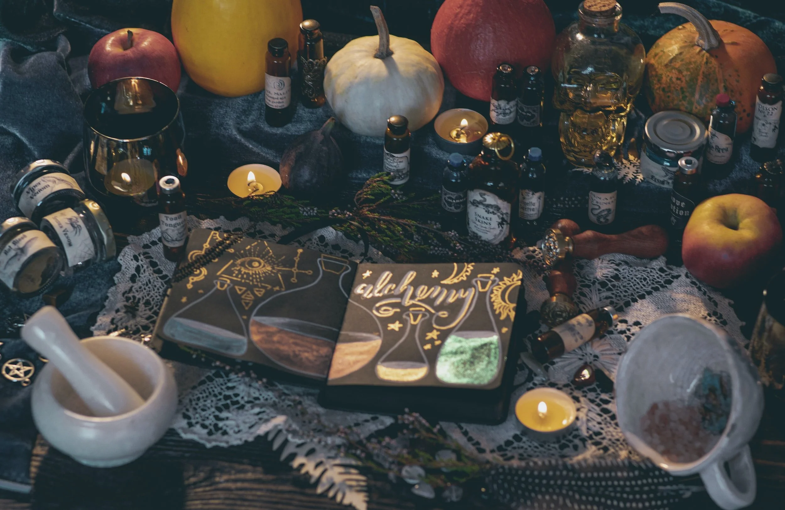 A ritual altar with candles, small bottles, pumpkins, apples, and a tarot card deck with the word 'alchemy' on it, arranged on a lace cloth.