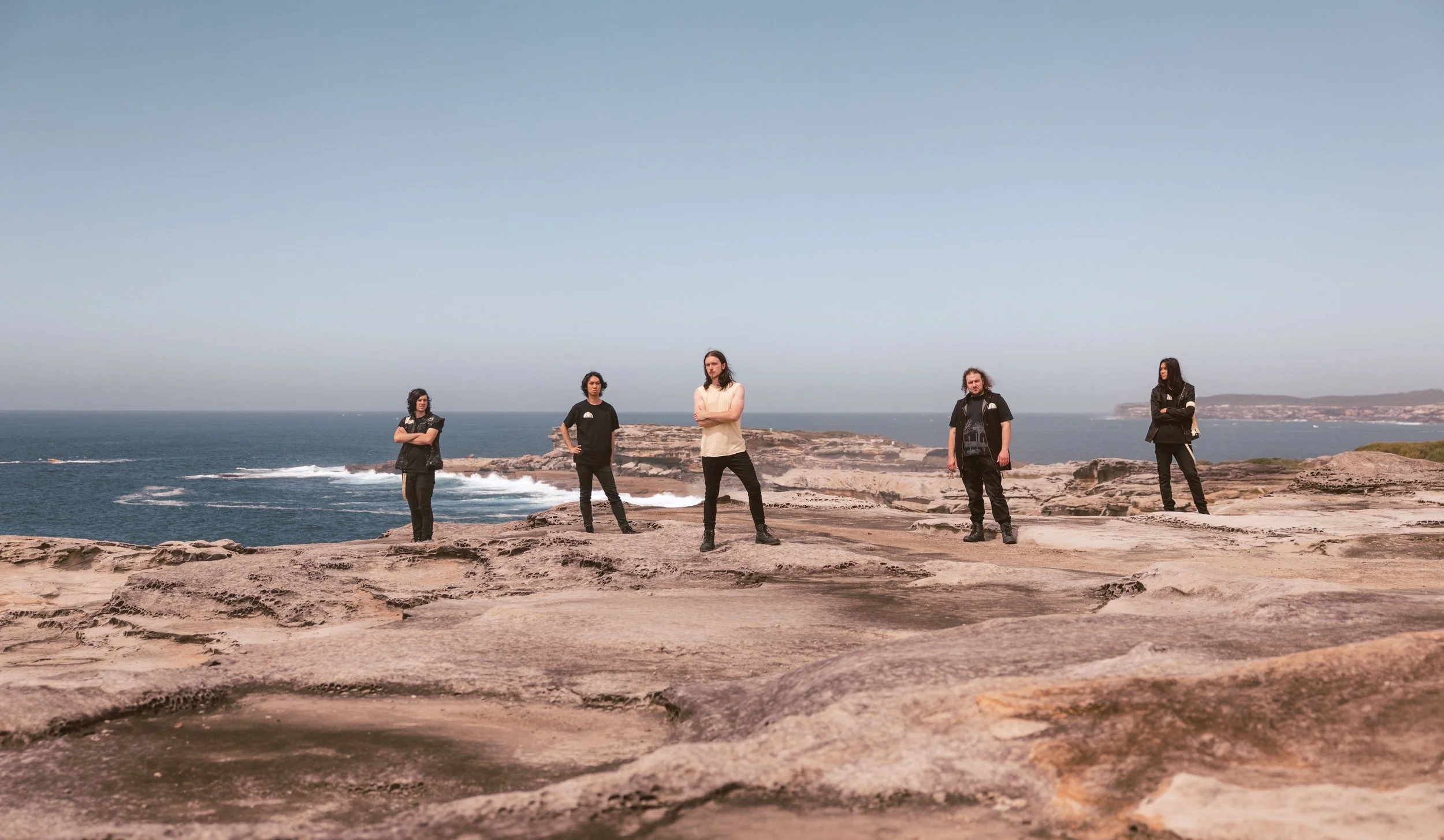 Five people standing on rocky coastal terrain with the ocean and cliffs in the background, under a clear blue sky.