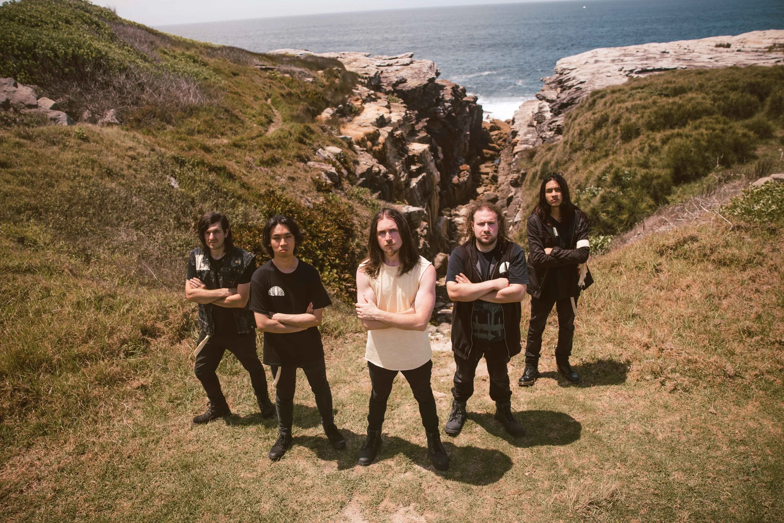 A group of five young men standing outdoors on grass near rocky cliffs with the ocean in the background. All have crossed arms and serious expressions.