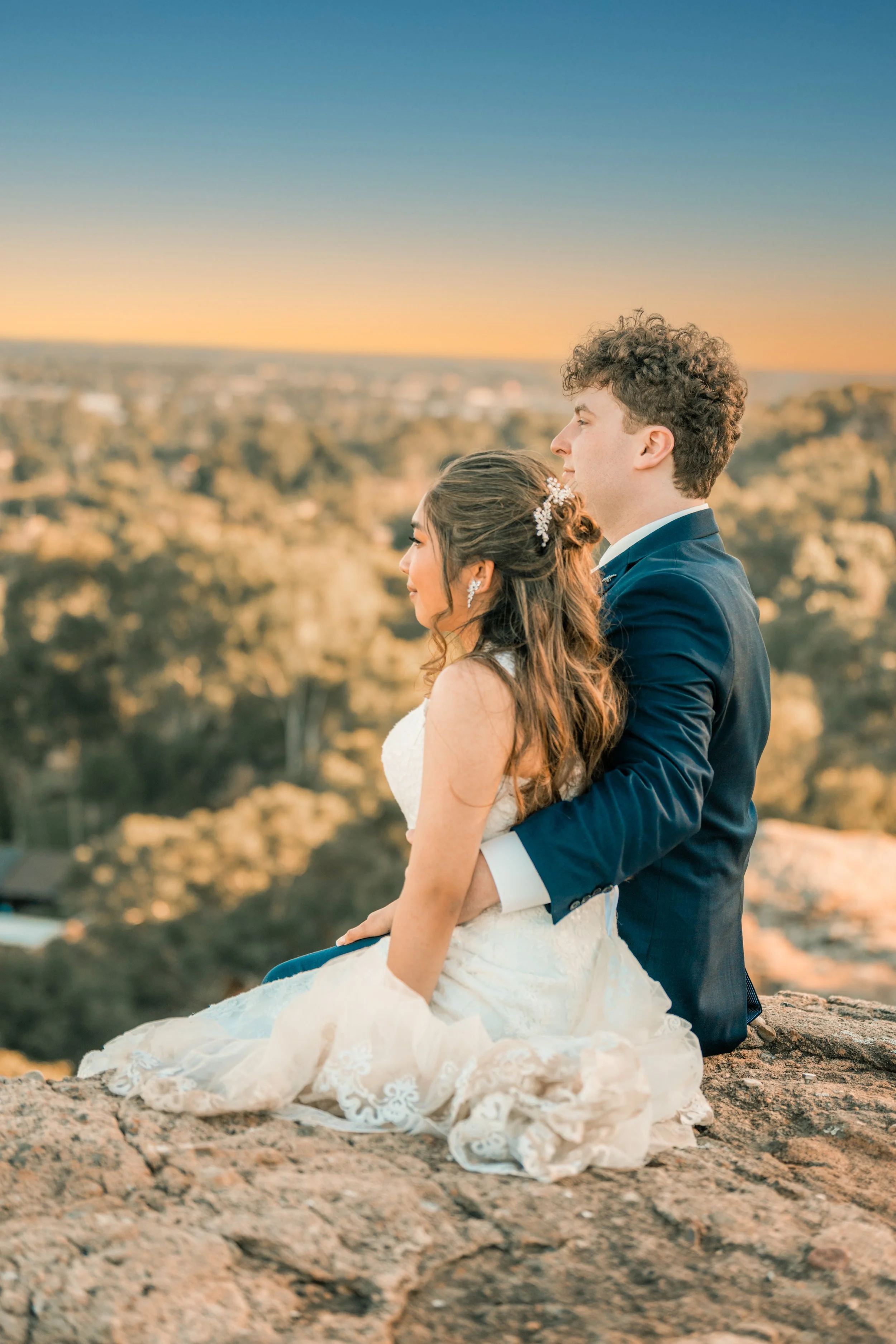 A newlywed couple sitting on a rocky outcrop, embracing each other and gazing at the sunset over a scenic landscape.