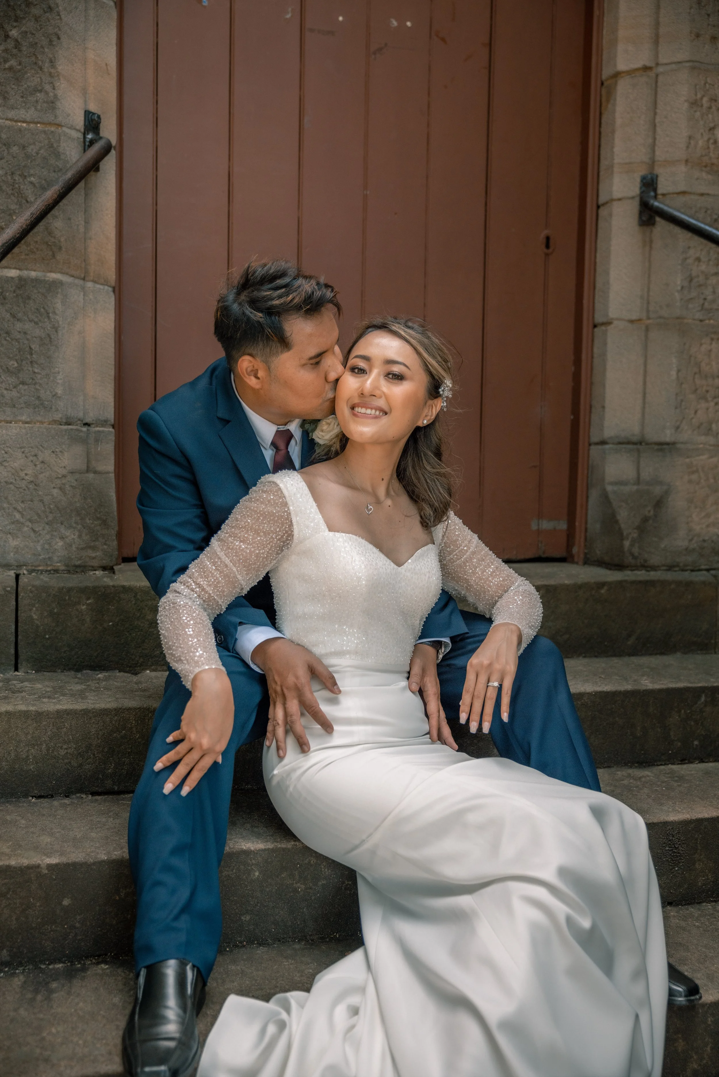 A newlywed couple sitting on stone steps outside of a building. The groom kisses the bride on the cheek while she smiles. The bride wears a white wedding dress with long sleeves, and the groom wears a navy suit with a white shirt and tie.