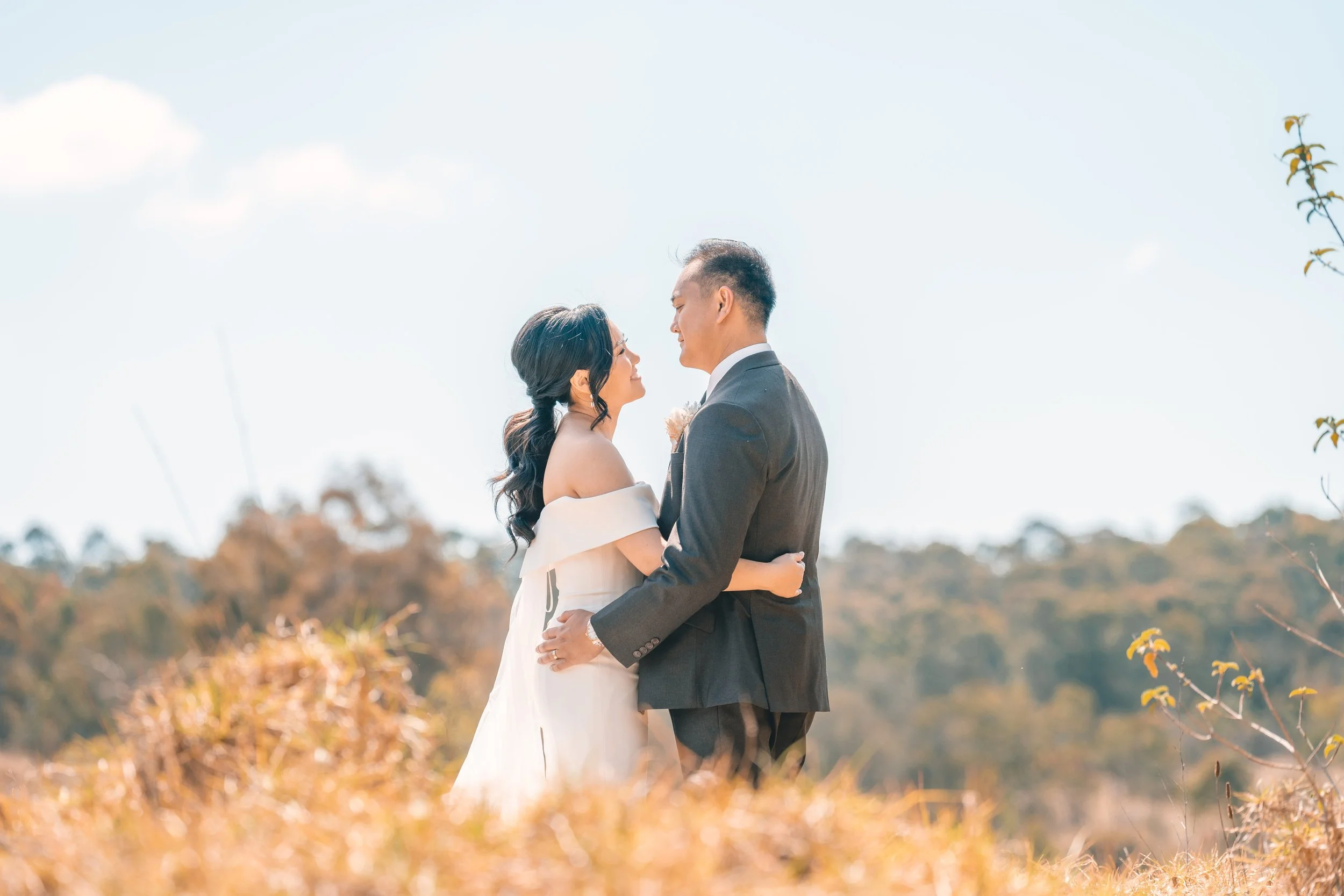 A bride and groom embracing outdoors in a scenic setting with trees in the background, on a bright, clear day.