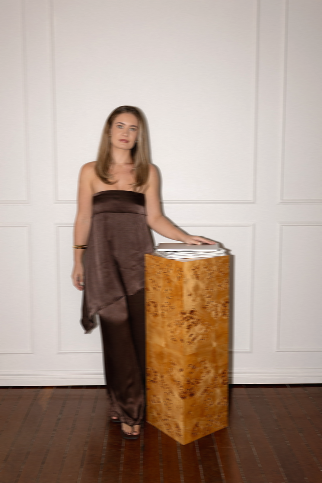 A woman standing next to a wooden pedestal with paperwork on top, against a white paneled wall, wearing a brown strapless outfit.
