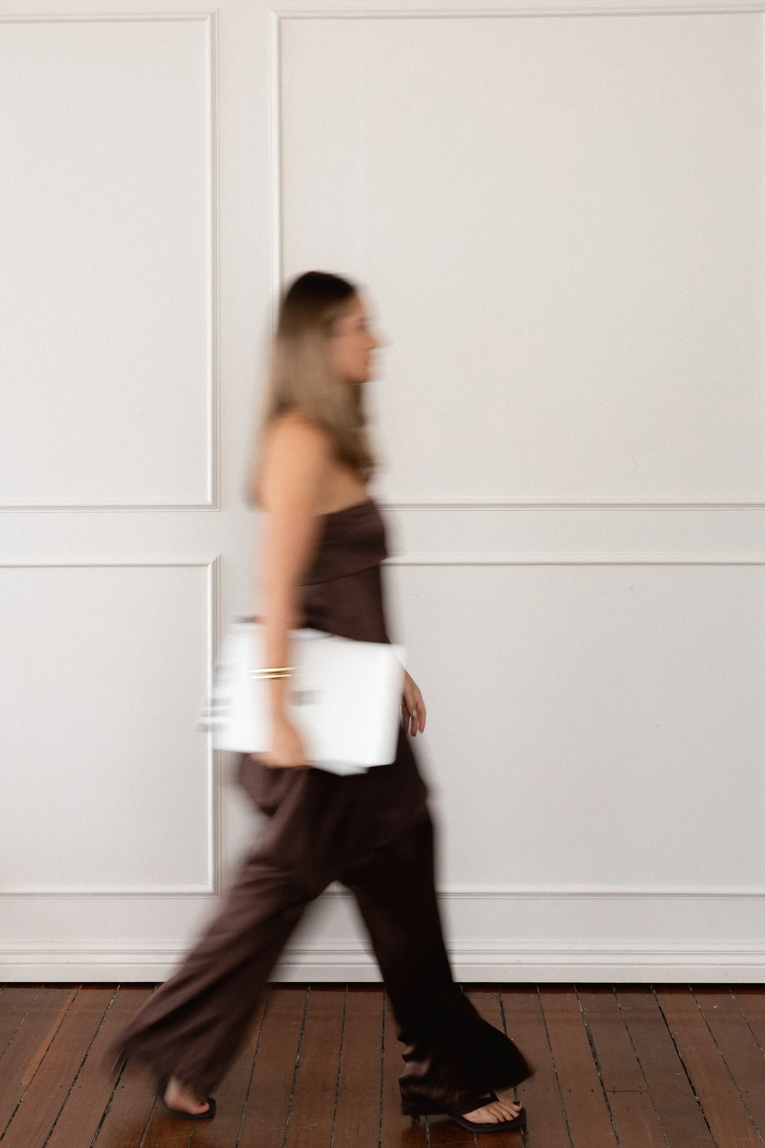 A woman walking barefoot on wooden floors in front of a white wall with rectangular paneling, wearing brown clothing and carrying a white tote bag.