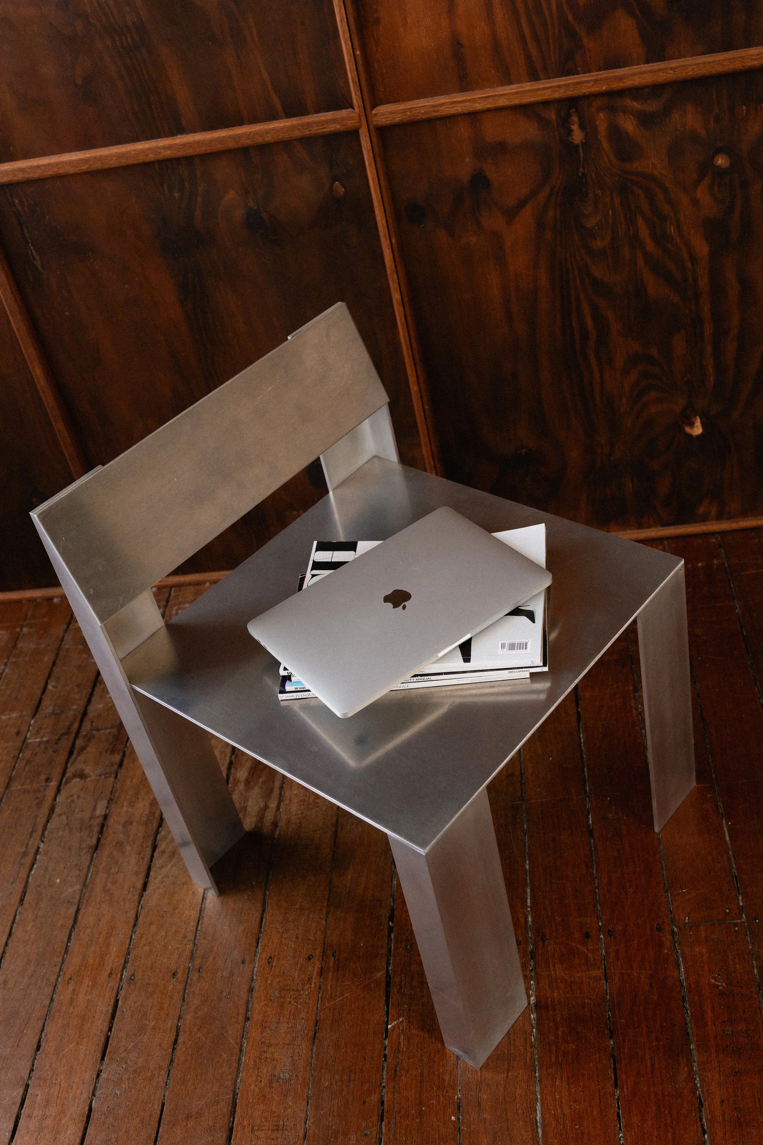 A silver chair with a flat seat and straight legs, placed on a wooden floor with a corner of a wooden cabinet or wall in the background. On the chair, there is a closed silver laptop and some magazines.