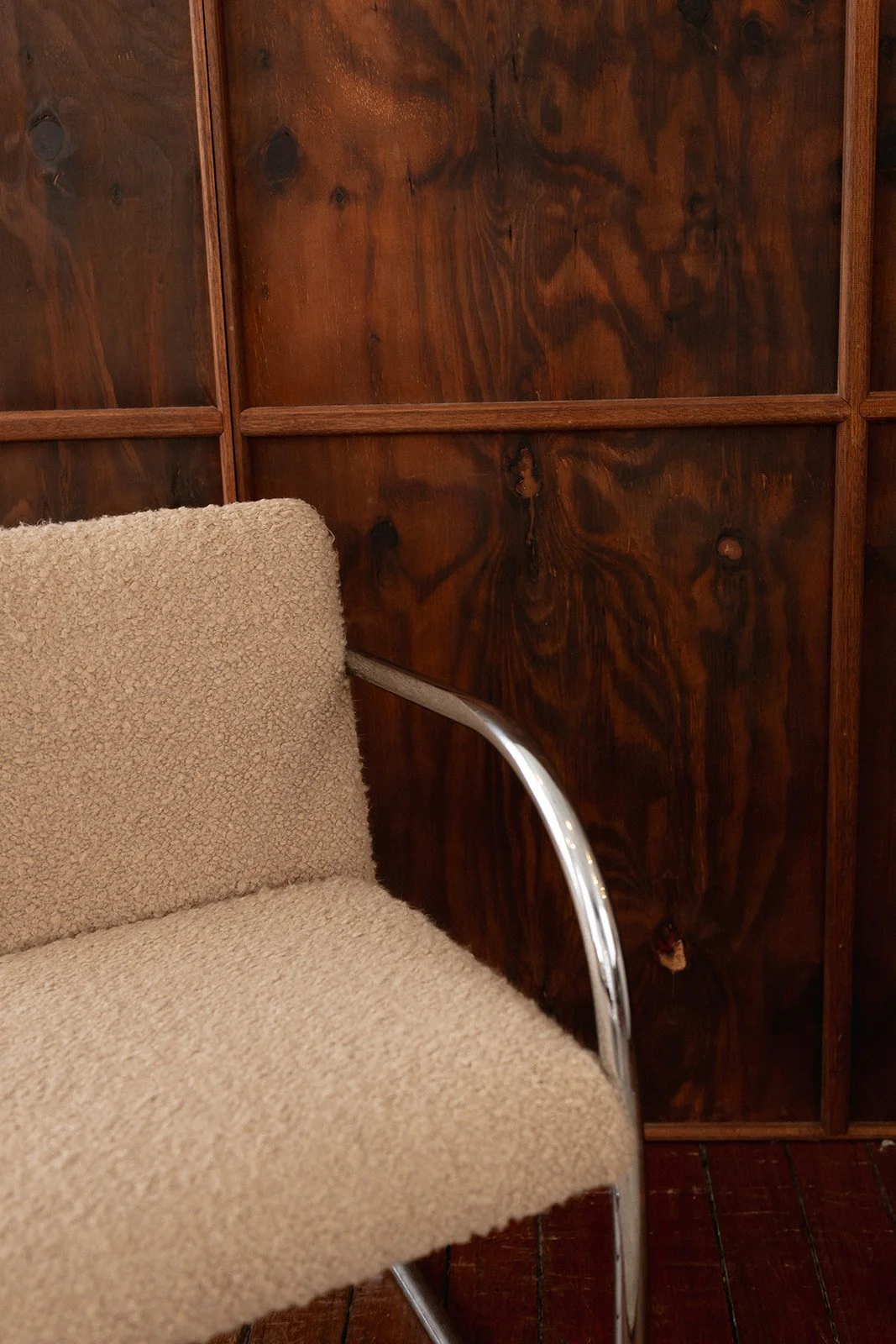 A beige textured upholstered chair with metallic armrest sits in front of a wooden wall made of dark stained planks and grid frames.