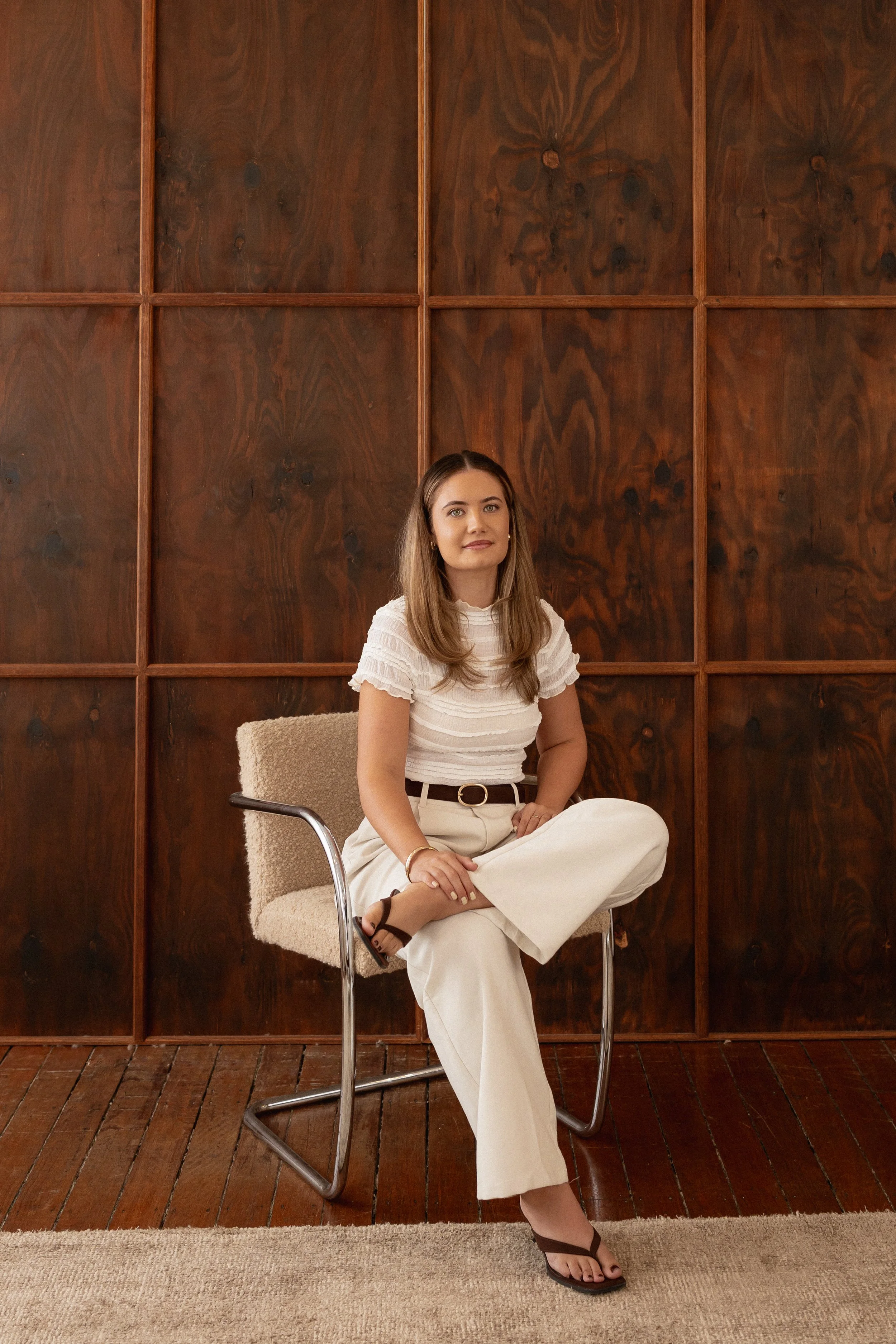 A woman sitting cross-legged on a chair with a wood-paneled wall background. She has long, light brown hair, wears a white top and wide-leg white pants with a belt, and black sandals. She has a neutral expression and is looking directly at the camera.