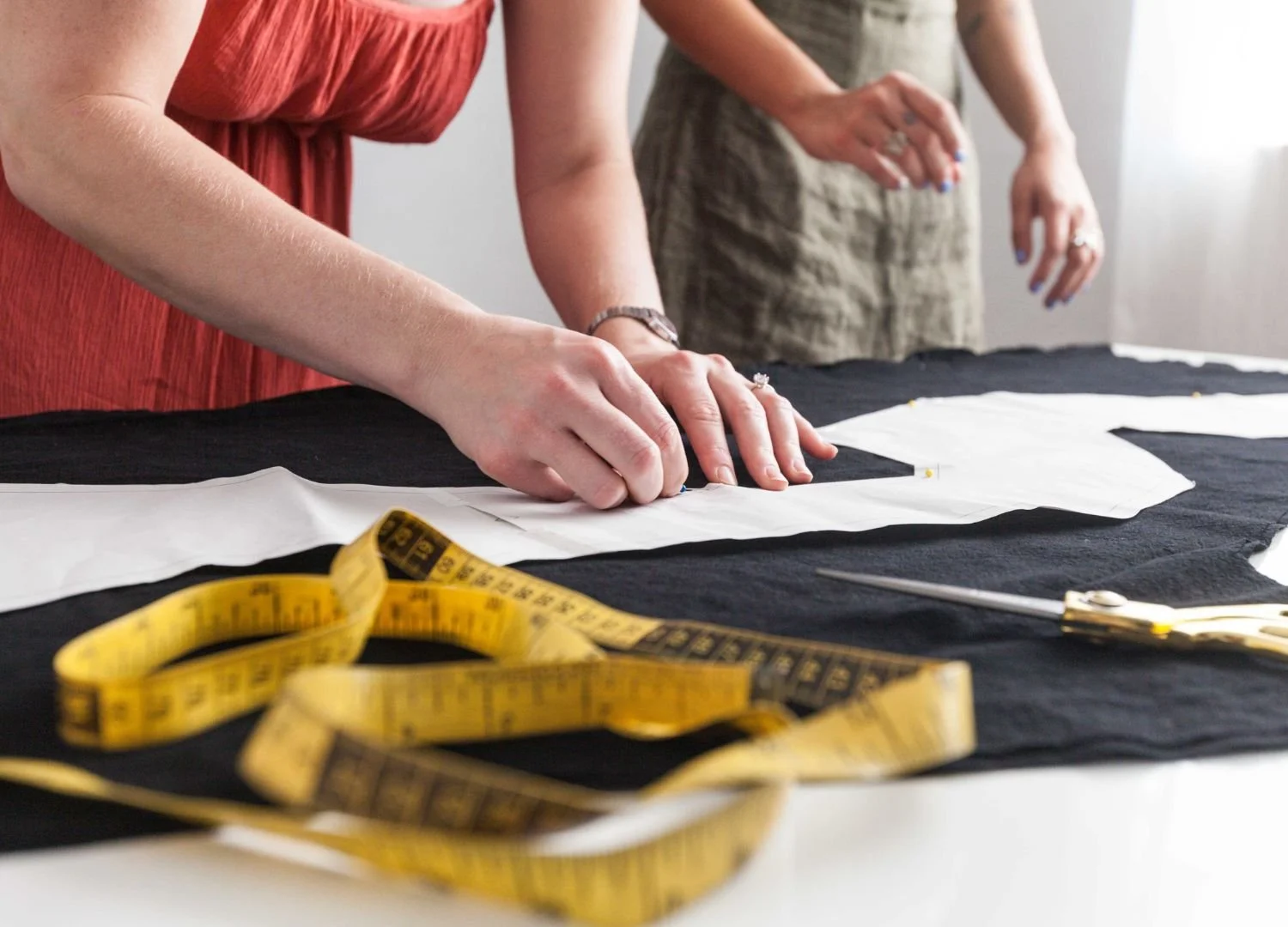 two people stand in front of table cutting out a pattern in black fabric with a tape measure in the foreground