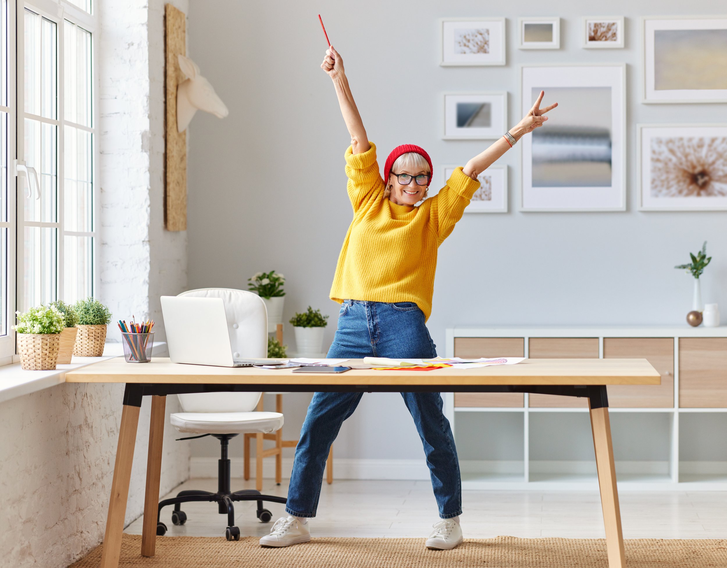 woman in yellow sweater, jeans and red beanie stands behind desk in well lit room, with her arms raised and smiling