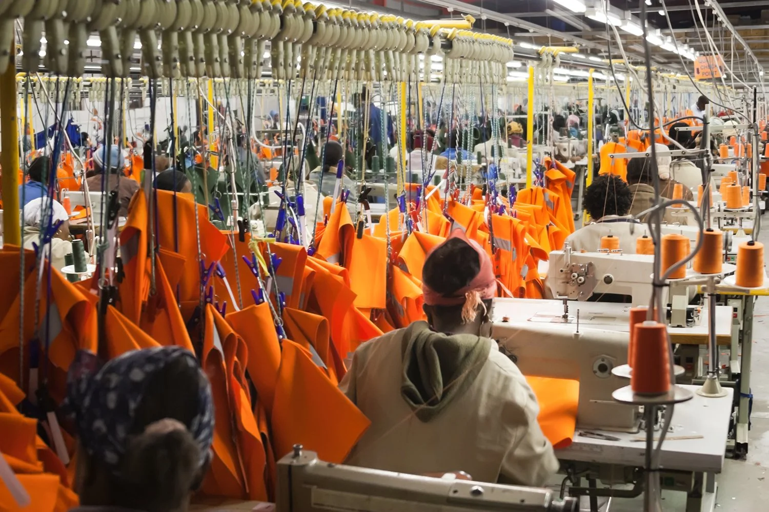 garment factory workers shown from behind lined up at their sewing machines. Orange projects hang from the center bar above them