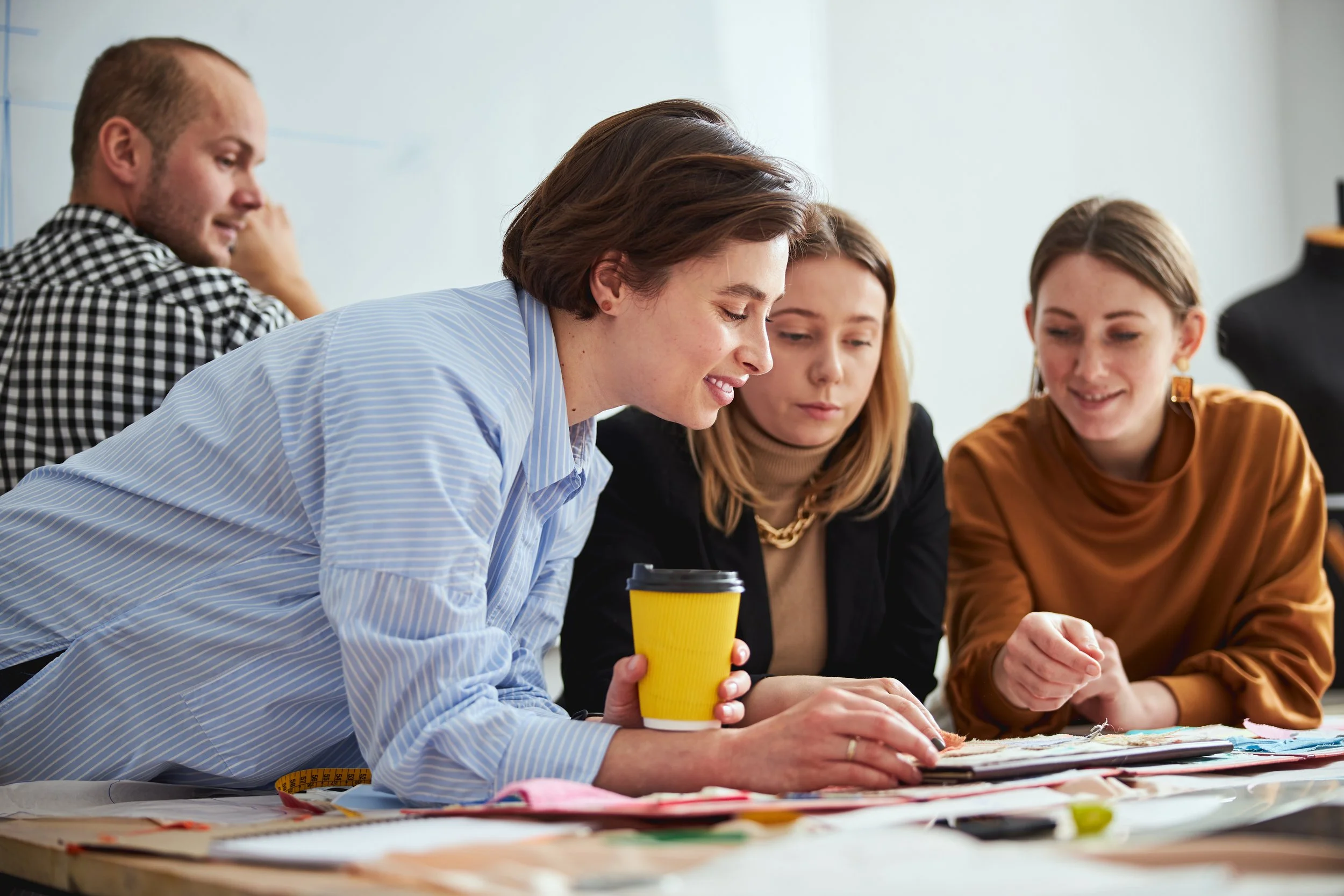 Fashion designer leans over table with a yellow coffee cup in hand and female pattern maker in a blaze and assistant look on at project details