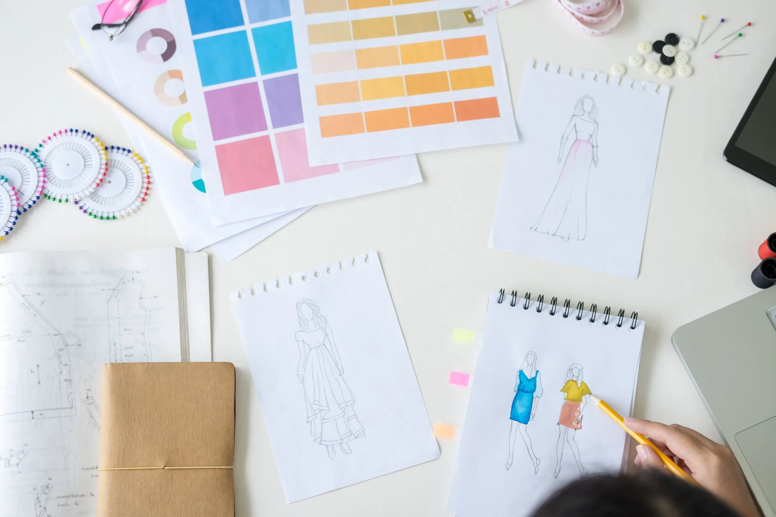 fashion designer sketching in notebook on a table surrounded by color swatches, colorful pin and diagrams