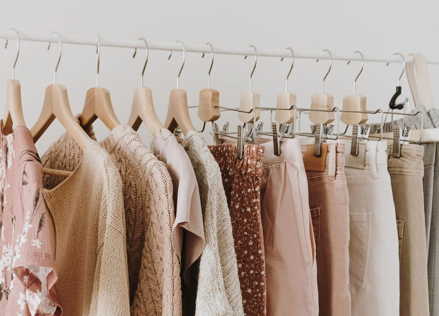 a closeup of a rack of clothing featuring pink and neutral colored sweaters, blouses and jeans
