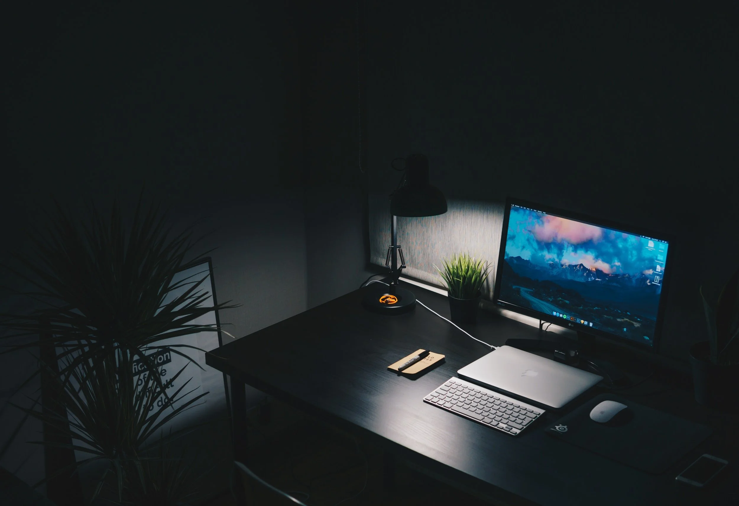 Dark organized desk with a computer monitor, laptop, keyboard, mouse, desk lamp, green plant, and a notepad with a pen, in a dimly lit room.