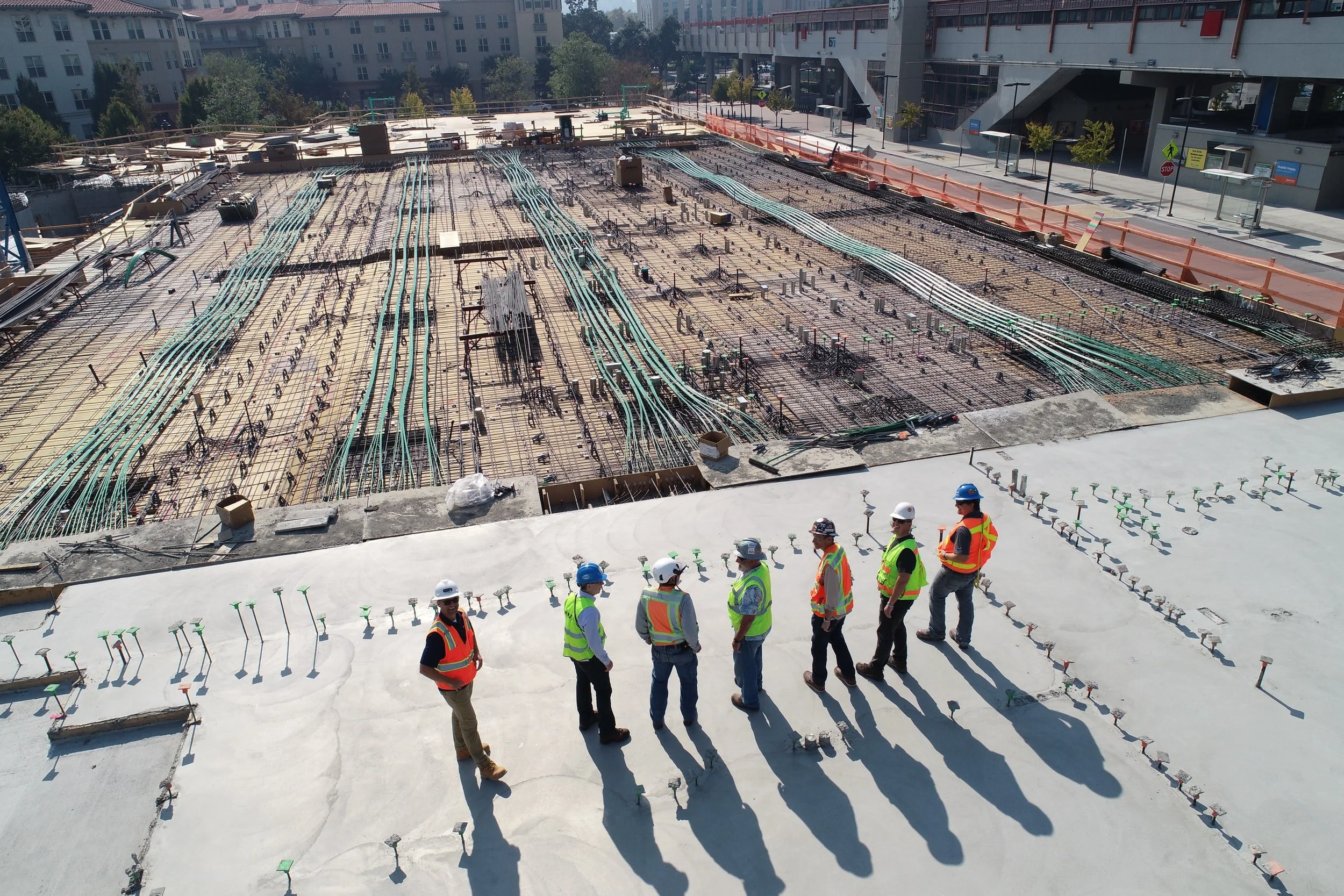 Group of construction workers and engineers wearing safety vests and helmets walking on a concrete roof at a construction site. In the background, there is a large open area with rebar and electrical wiring for a building's foundation or floor.
