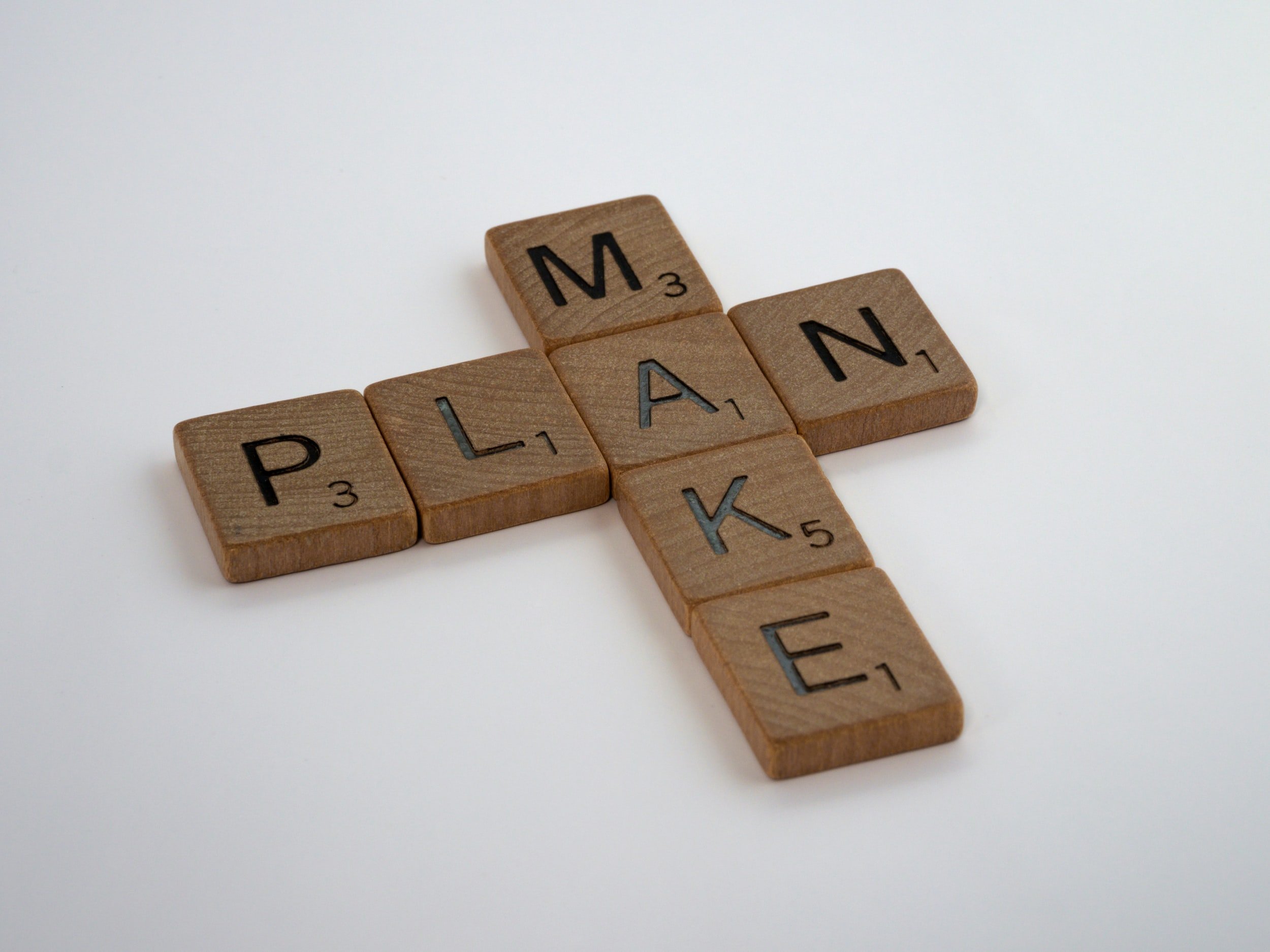 Scrabble tiles arranged in the shape of a cross spelling the words 'MAKE' and 'PLAN' on a white background.