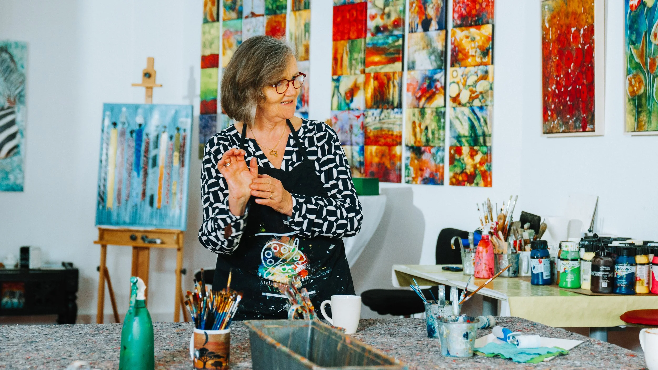 Frau in Atelier mit Malutensilien und bunten Gemälden an der Wand.