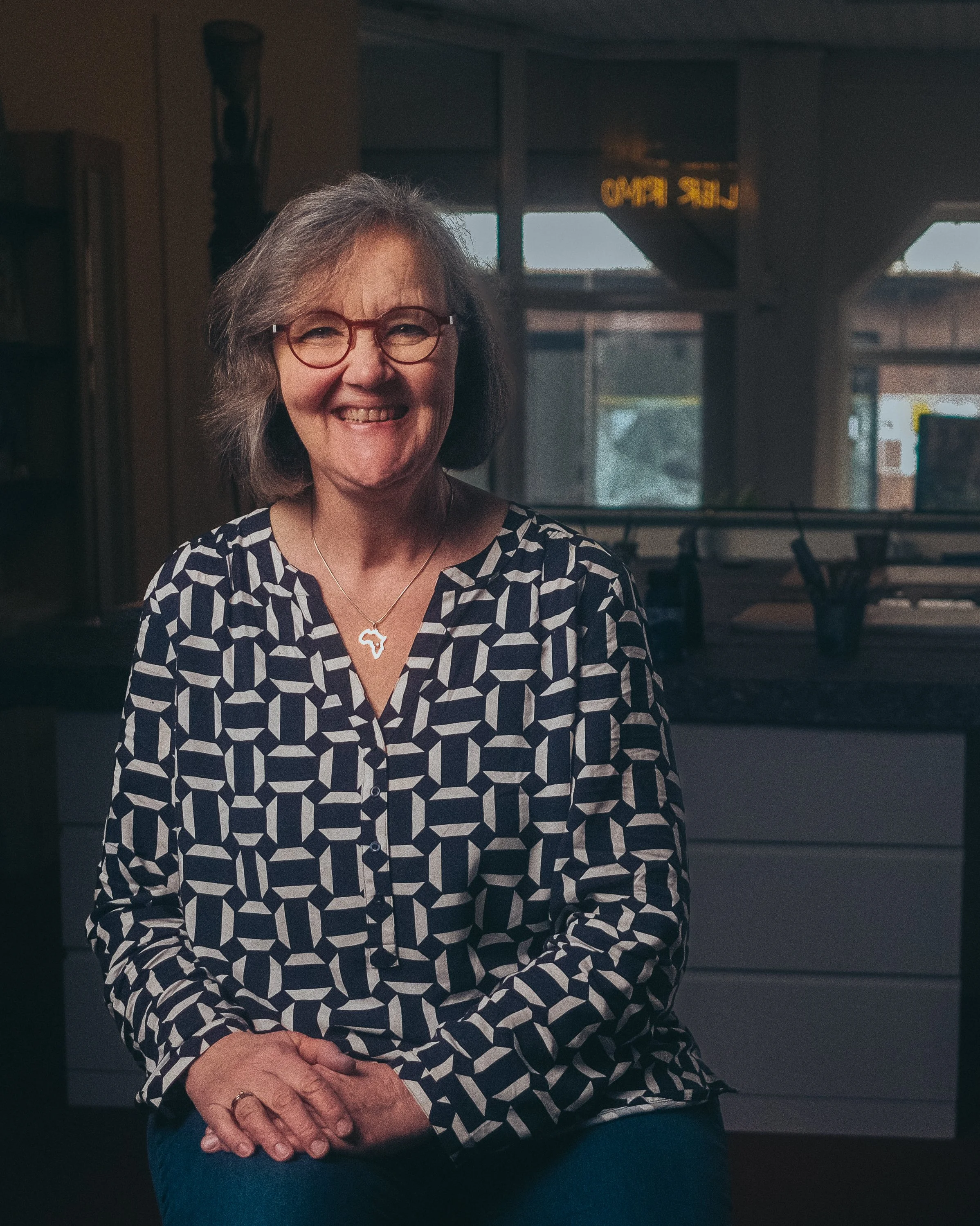 Frau mit Brille und geometrisch gemusterter Bluse lächelt in die Kamera, Innenaufnahme in einem Raum mit dunklem Hintergrund.