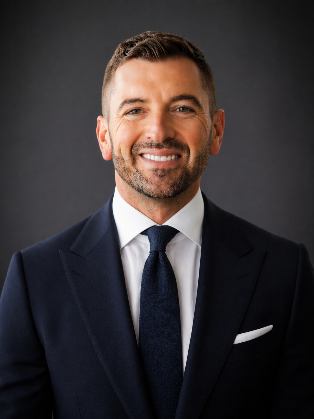 Portrait of a smiling man in a dark suit, white shirt, and dark tie, against a dark background.