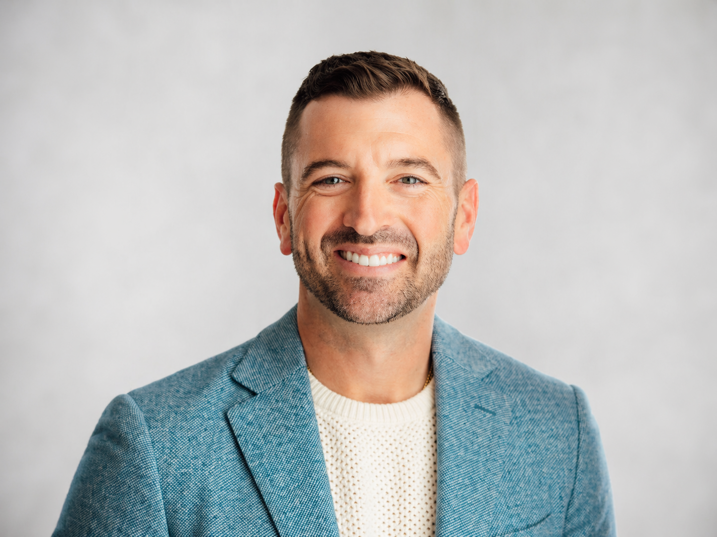 Portrait of a smiling man with short brown hair and a beard, wearing a light blue blazer over a cream sweater, against a plain white background.