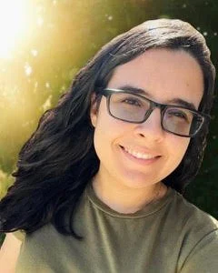 A smiling woman with long dark hair, glasses, and a green shirt outdoors with sunlight and trees in the background.