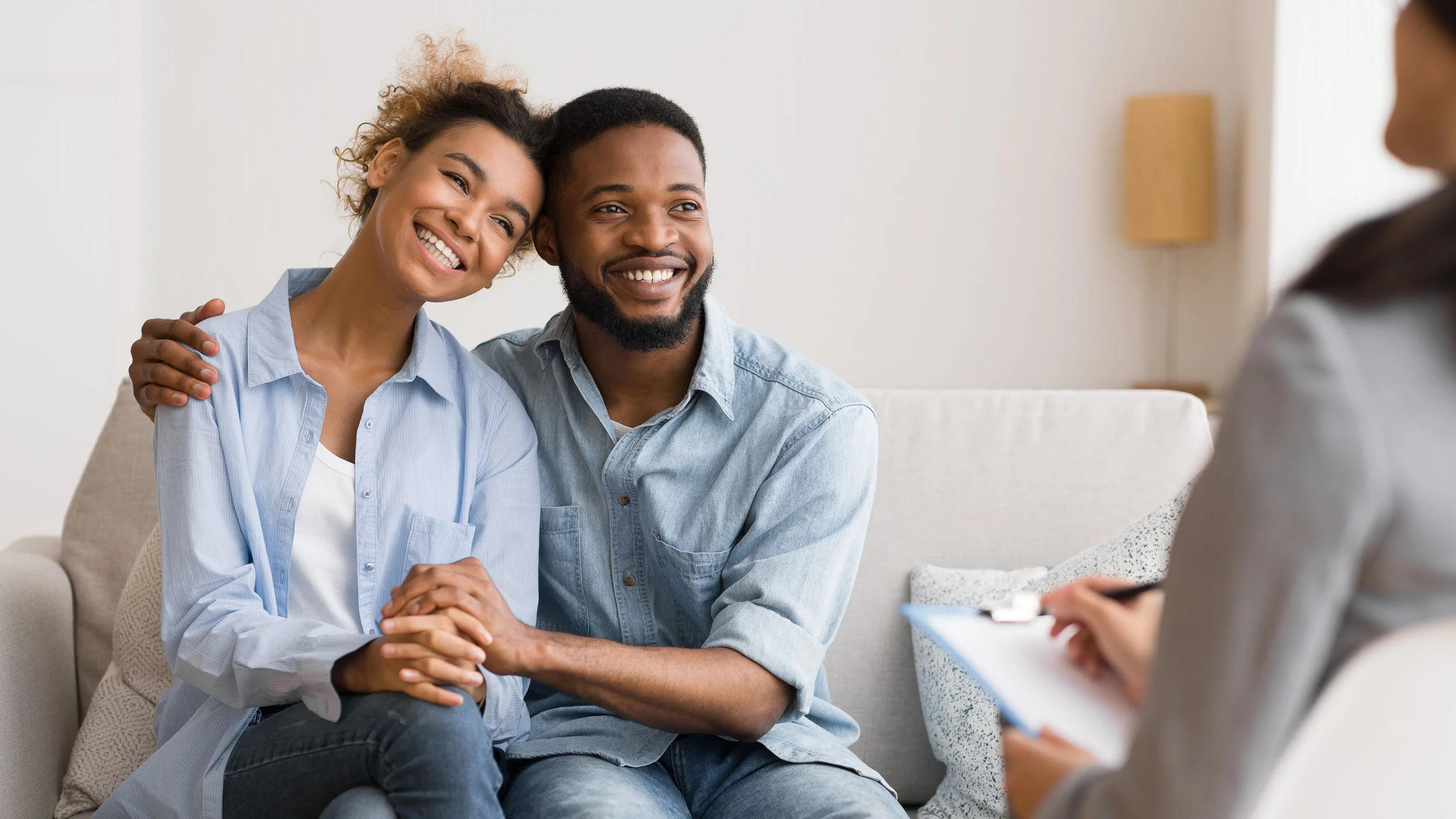 Couple sitting close together on a couch during a therapy session, smiling as they talk to a counselor holding a clipboard — representing the compassionate relationship support offered by Let's Talk 4 Health’s online counseling services.