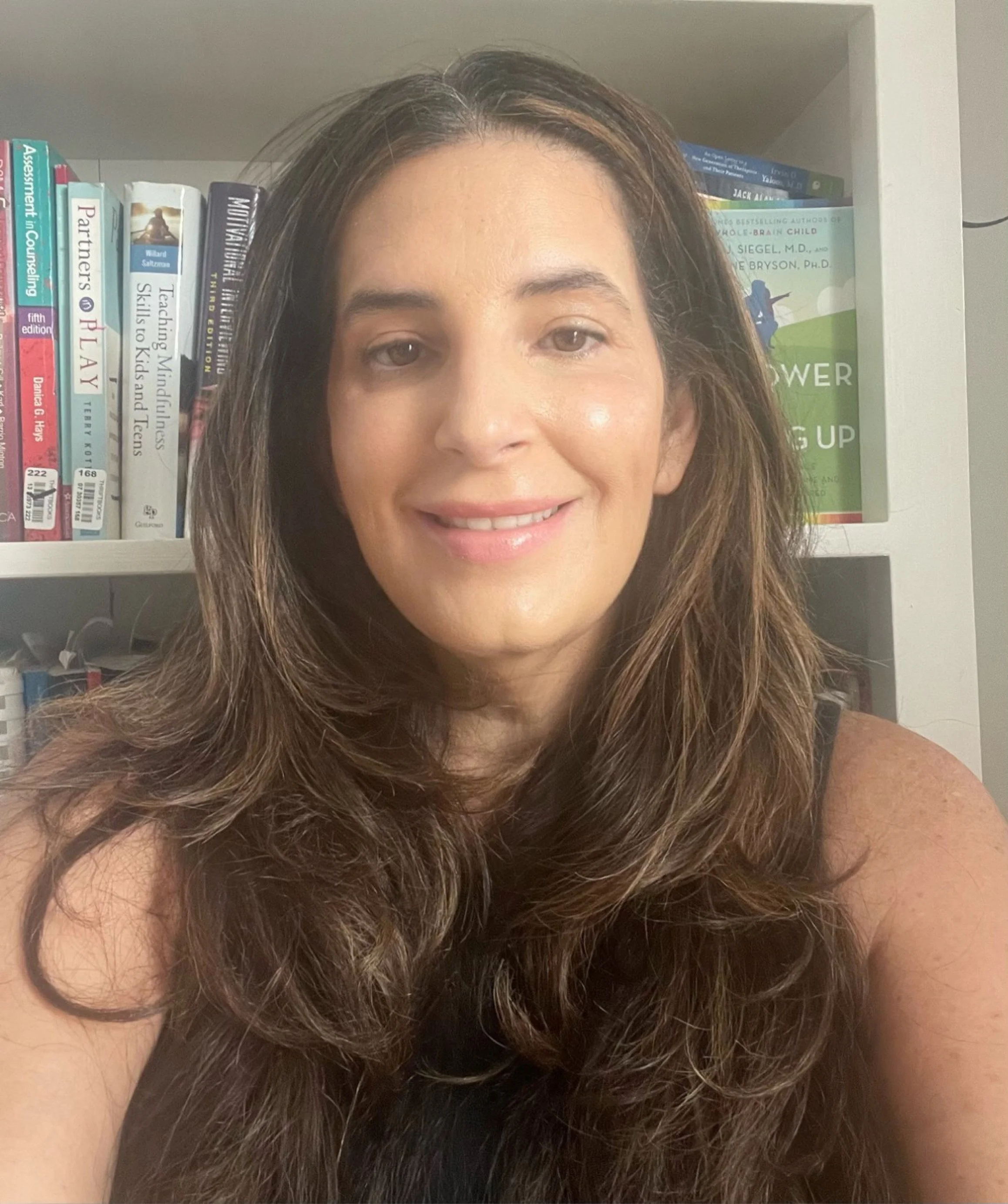 A woman with long, wavy brown hair smiling at the camera, with a bookshelf in the background.