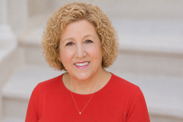 A smiling middle-aged woman with short, curly blond hair, wearing a red top with floral patterns.