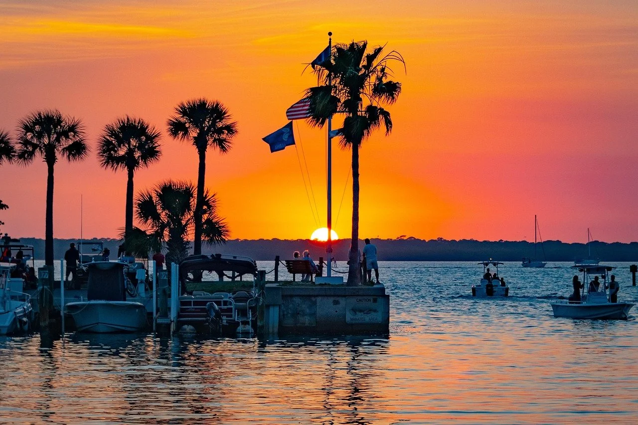 Sunset over a marina with boats, palm trees, and people by the water, with a vibrant orange and pink sky.