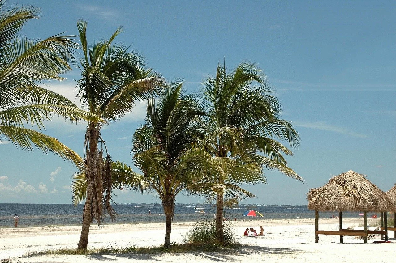 A tropical beach with palm trees, a thatched hut, and a few people relaxing under a colorful umbrella in the sand.