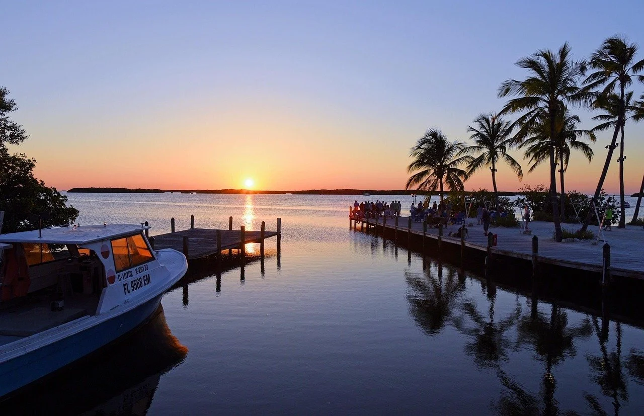 Sunset over a calm body of water with a dock, a boat, and people gathered on a pier and around palm trees, creating a tropical, relaxing scene.