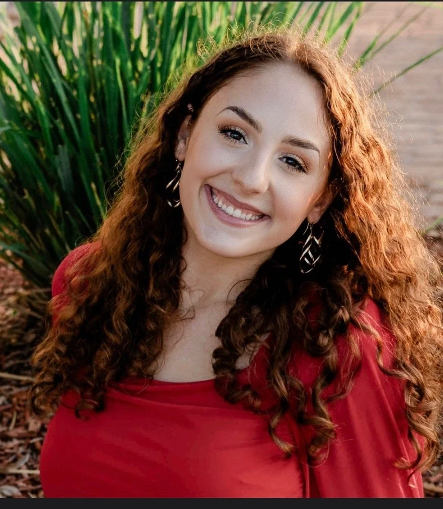A young woman with long, curly red hair and light skin, smiling outdoors, wearing a red top with large silver earrings, standing in front of green plants and a body of water.