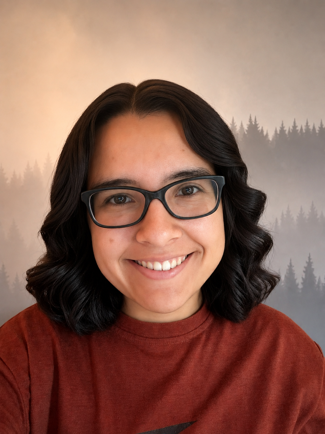 A smiling woman with long dark hair, glasses, and a green shirt outdoors with sunlight and trees in the background.