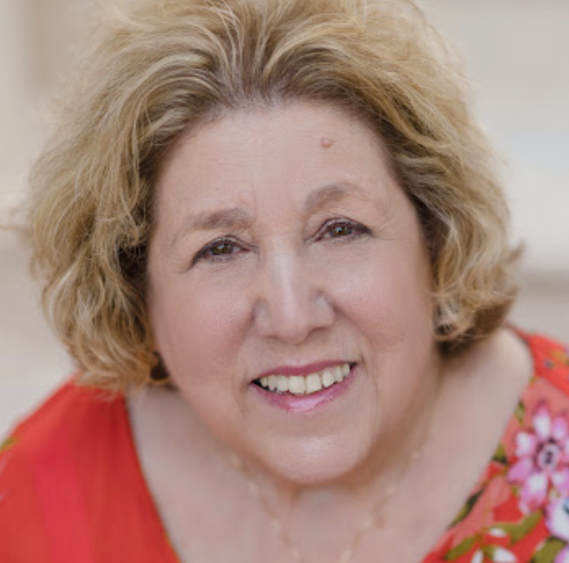 A smiling middle-aged woman with short, curly blond hair, wearing a red top with floral patterns.