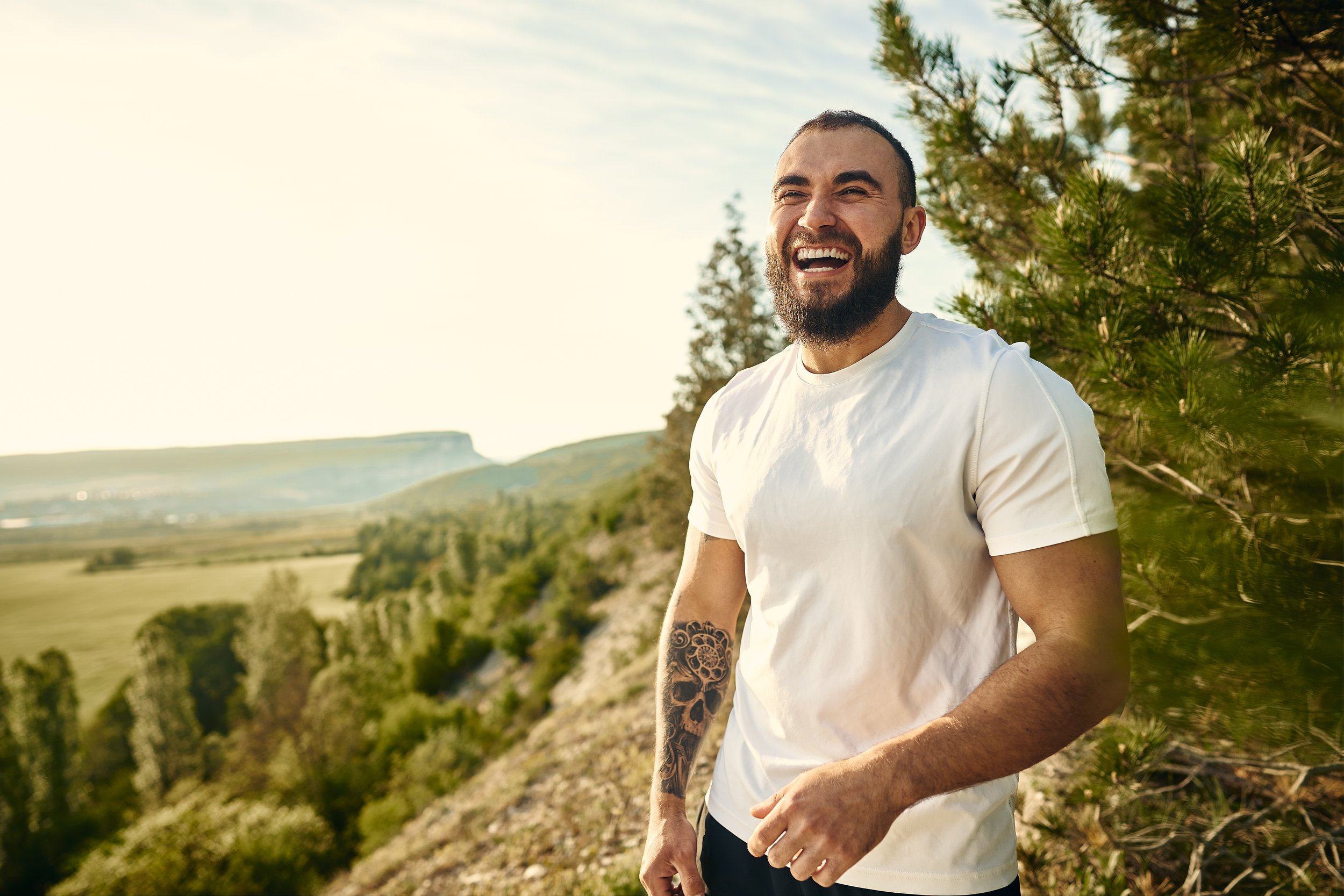 A smiling man with a beard and a tattoo on his arm standing outdoors near trees, with hills and open fields in the background — reflecting confidence, individuality, and the personal growth supported by Let's Talk 4 Health’s mental health services.