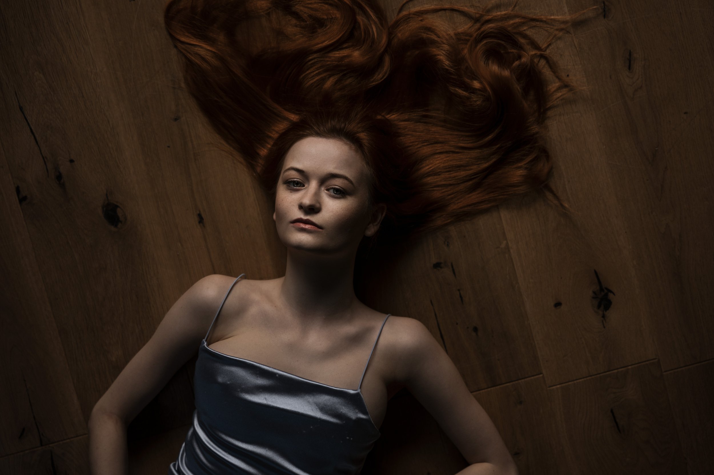 Junge Frau liegt auf Holzboden, lange rotes Haar ausgebreitet um den Kopf, trägt silbernes Satin-Nachtkleid, blickt direkt in Kamera.